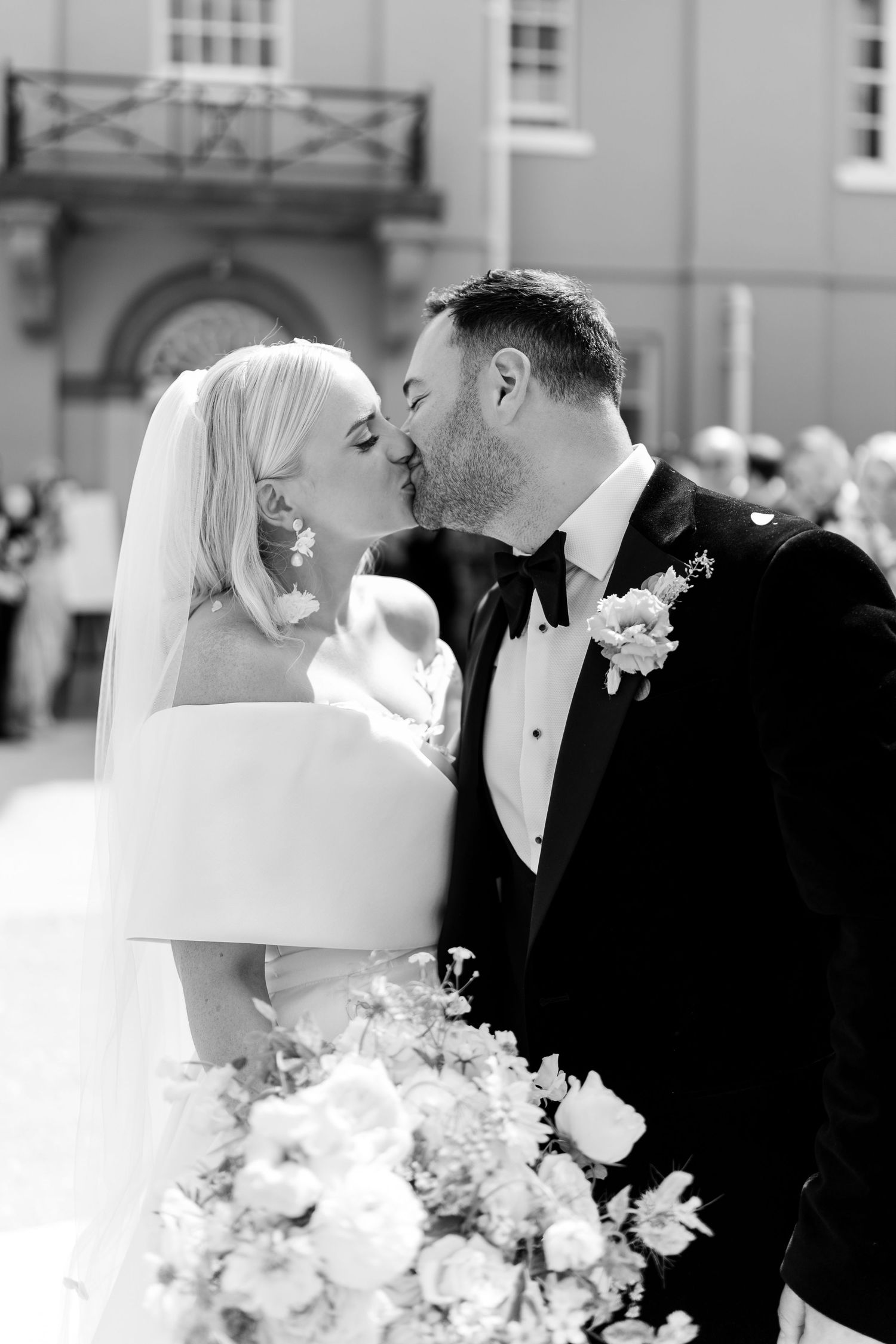 Newlyweds share first kiss as bride holds lush floral bouquet outside historic building.