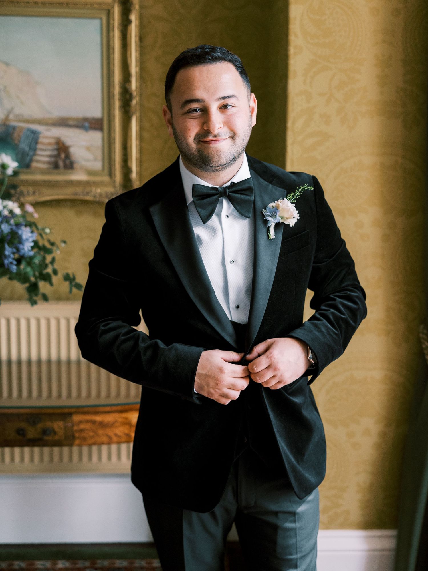 Groom in black velvet tuxedo with bow tie and floral boutonniere posing indoors.