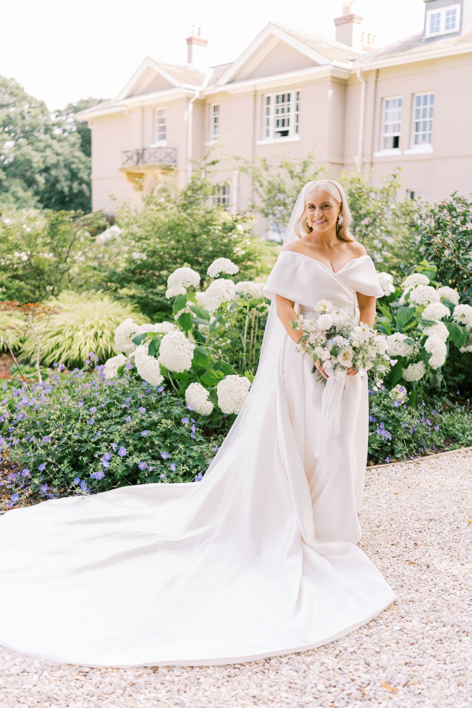 Bride in off-shoulder white gown with long train holds bouquet among white hydrangeas outside elegant pink manor.