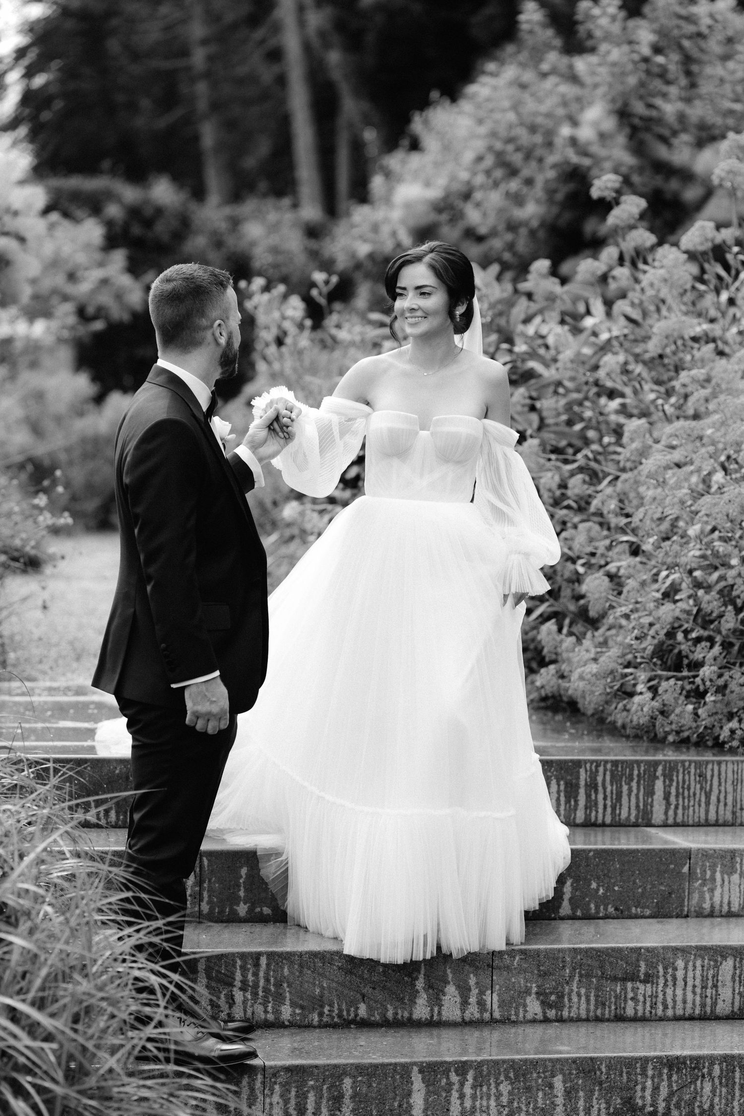 Black and white photo of smiling bride in off-shoulder gown holding bouquet with groom on garden steps.