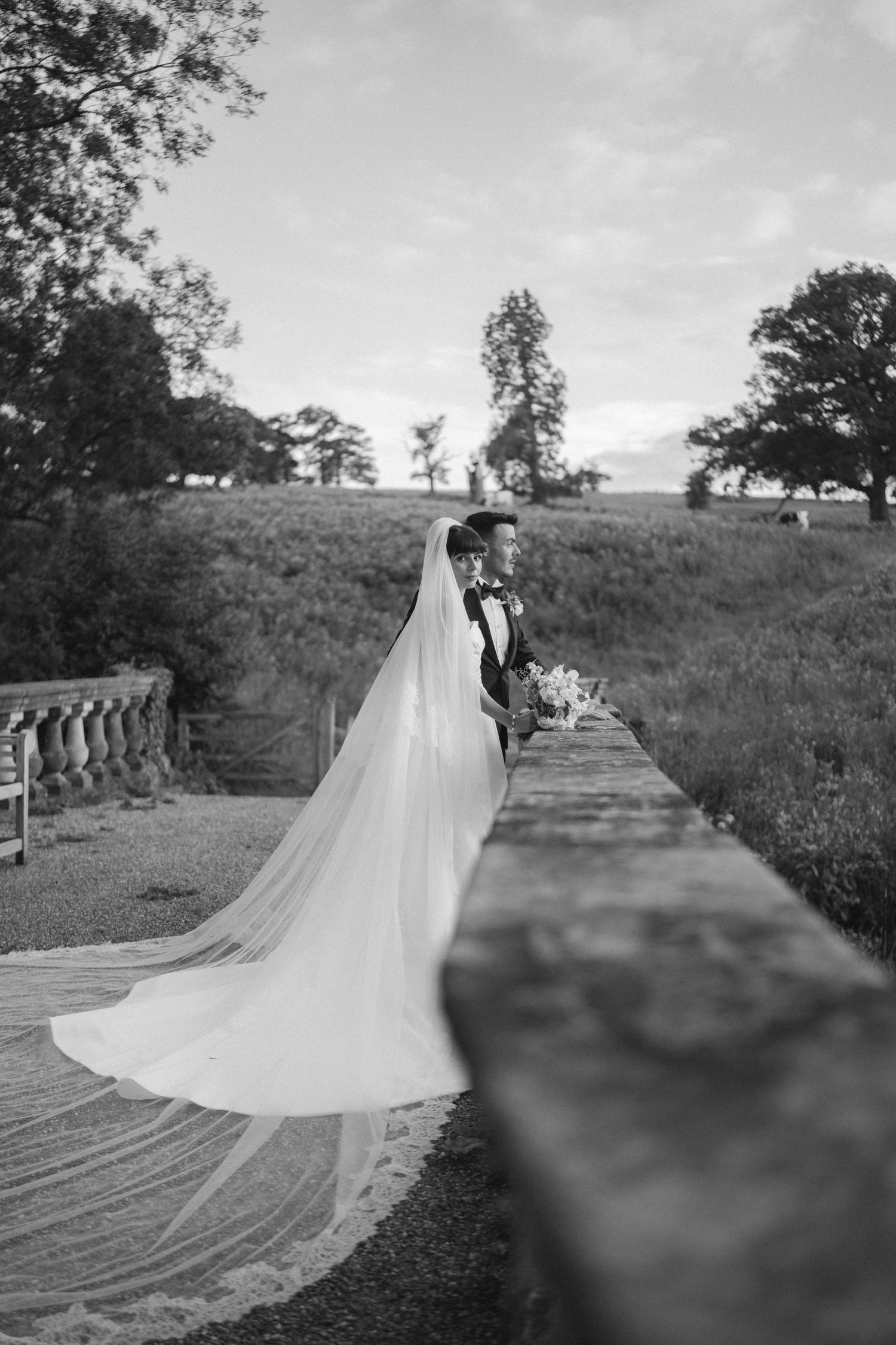 Black and white wedding photo of bride and groom standing on stone terrace, cathedral veil flowing, lush greenery backdrop.