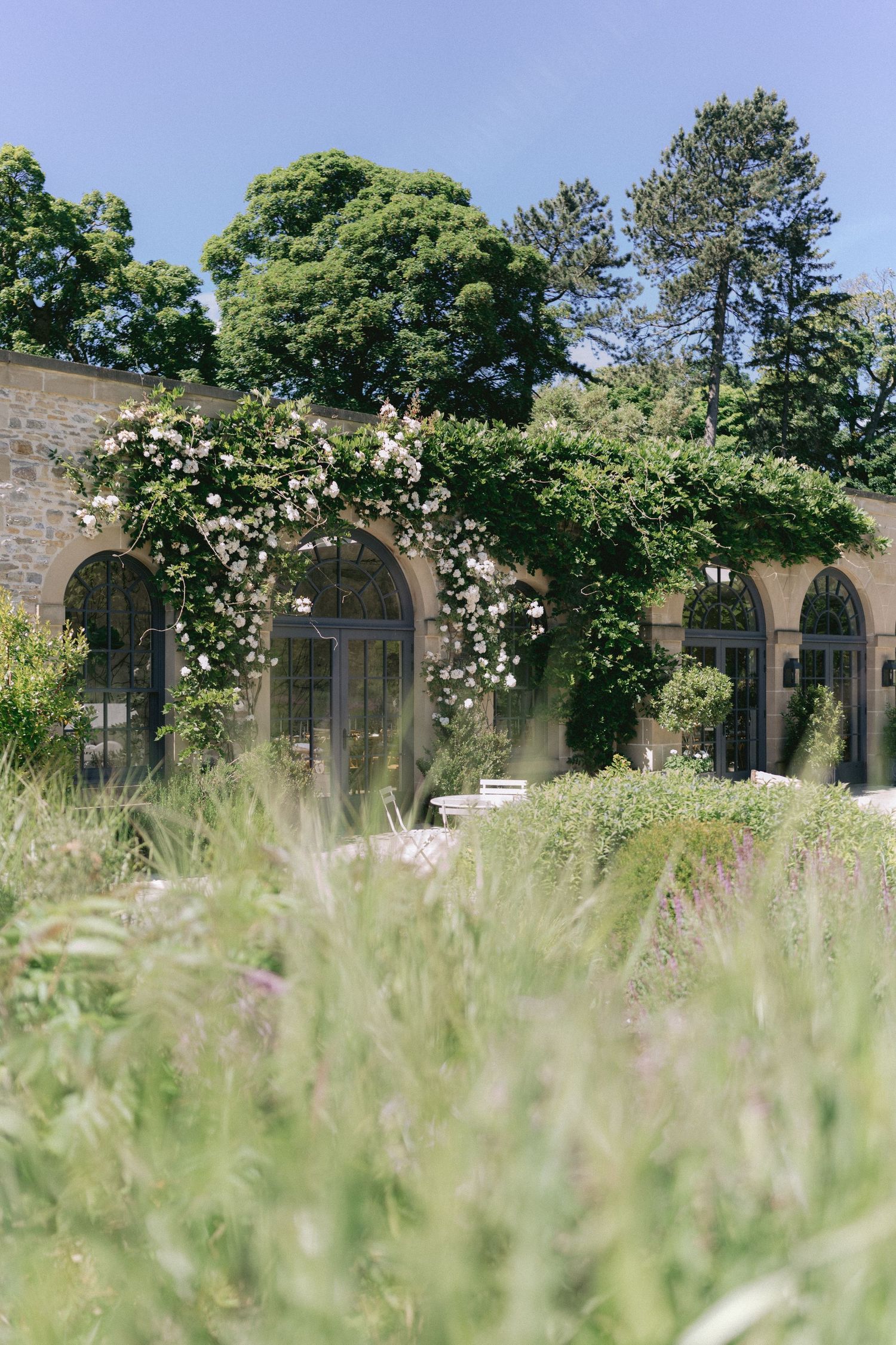 Stone building with arched windows covered in climbing roses, surrounded by lush green gardens under a clear blue sky.