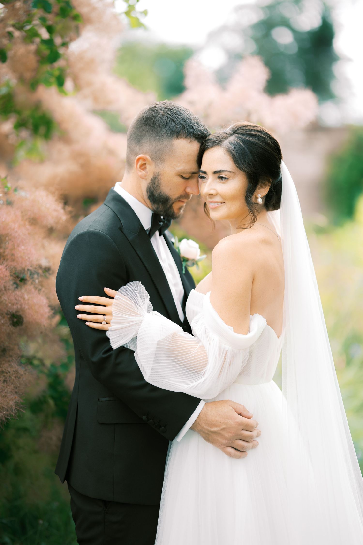 Bride and groom sharing an intimate forehead touch surrounded by soft pink blooms in a romantic garden wedding portrait.