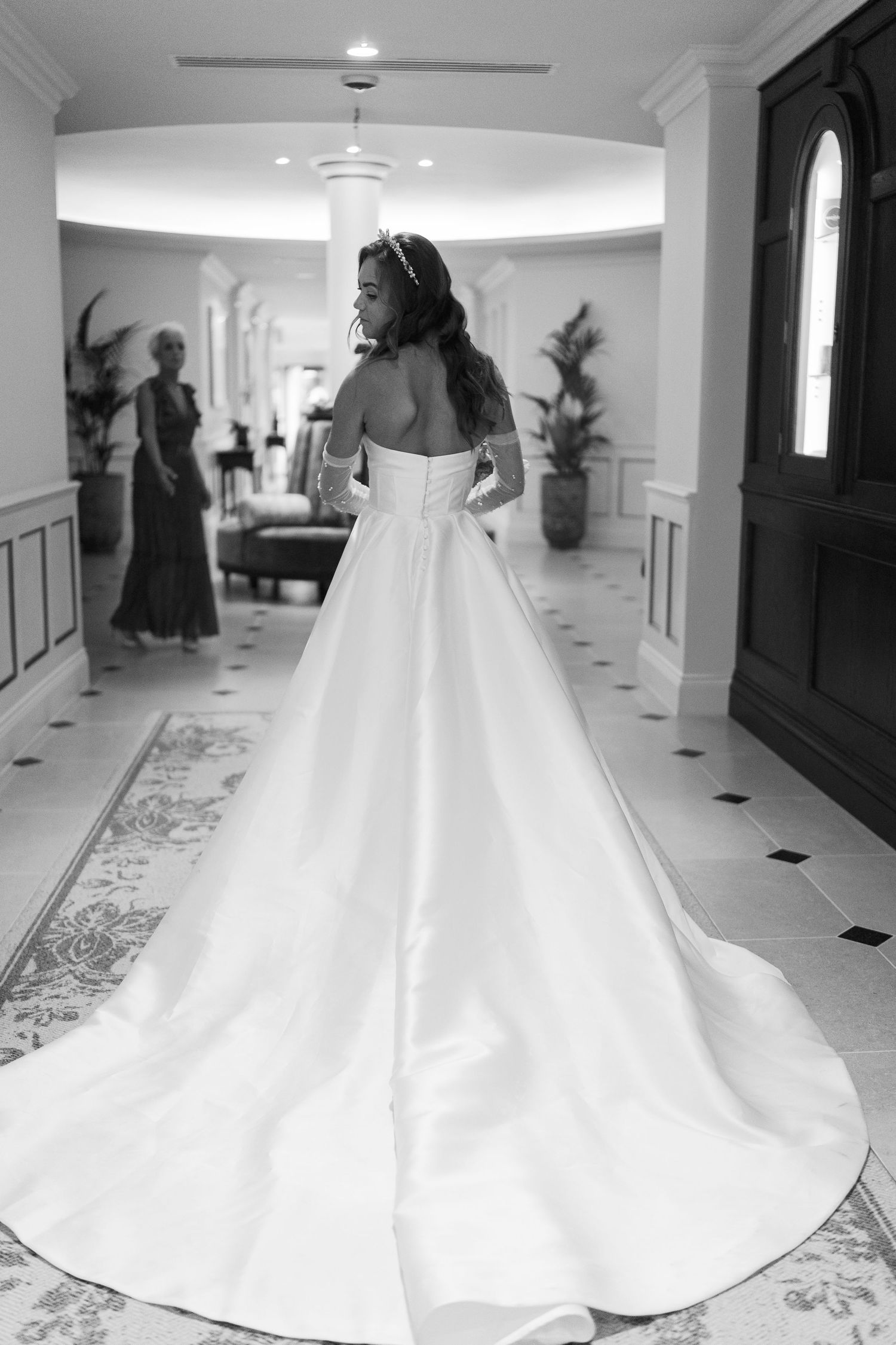 Bride in elegant strapless ball gown with long train viewed from behind in grand hotel hallway.