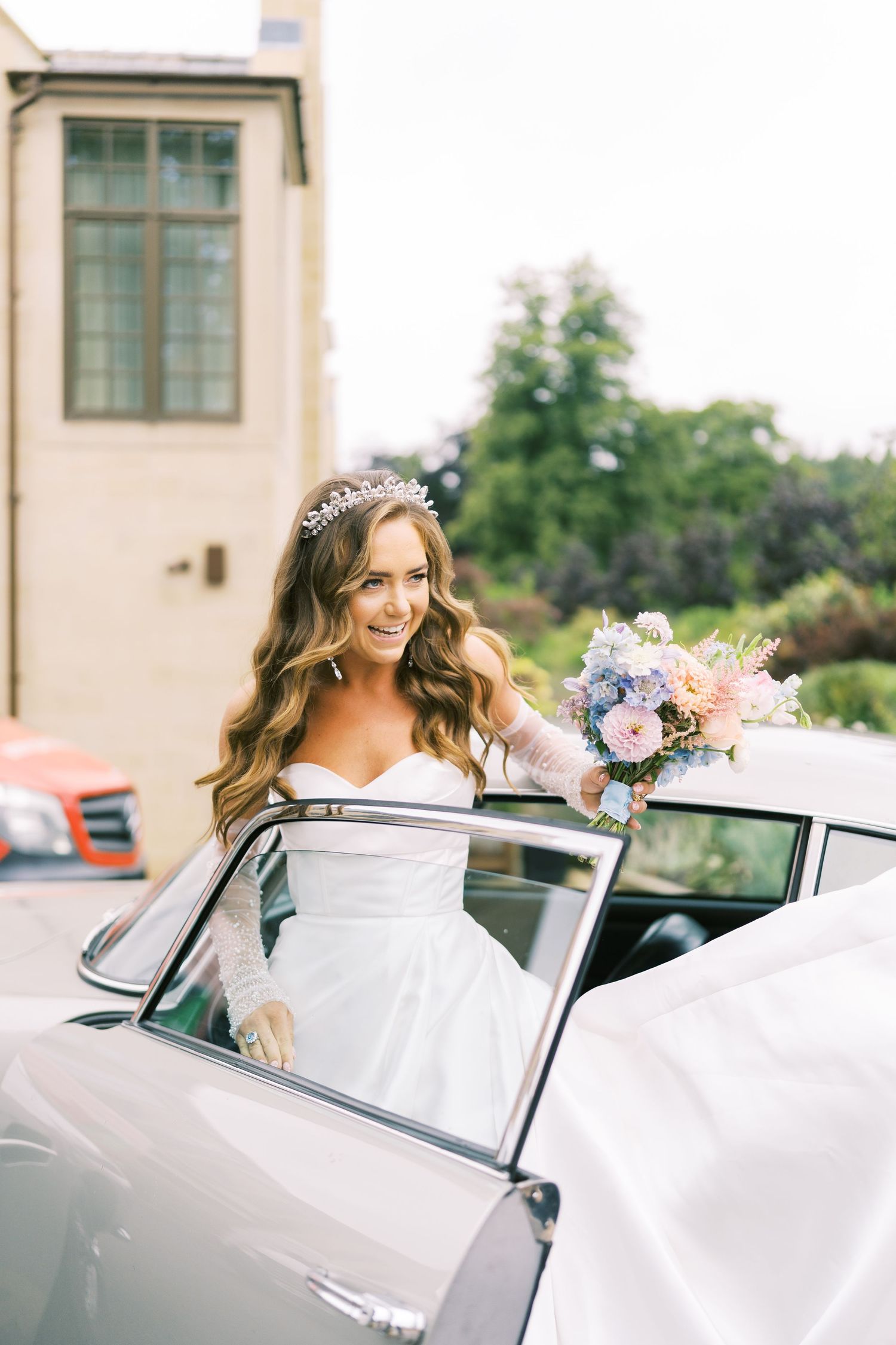 Bride in white gown and tiara holds floral bouquet stepping out of vintage car on wedding day.