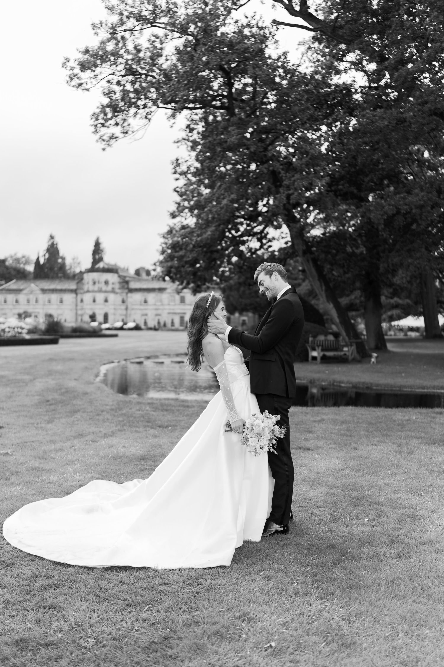 Black and white photo of bride and groom embracing on a lawn with a grand estate and trees in the background.