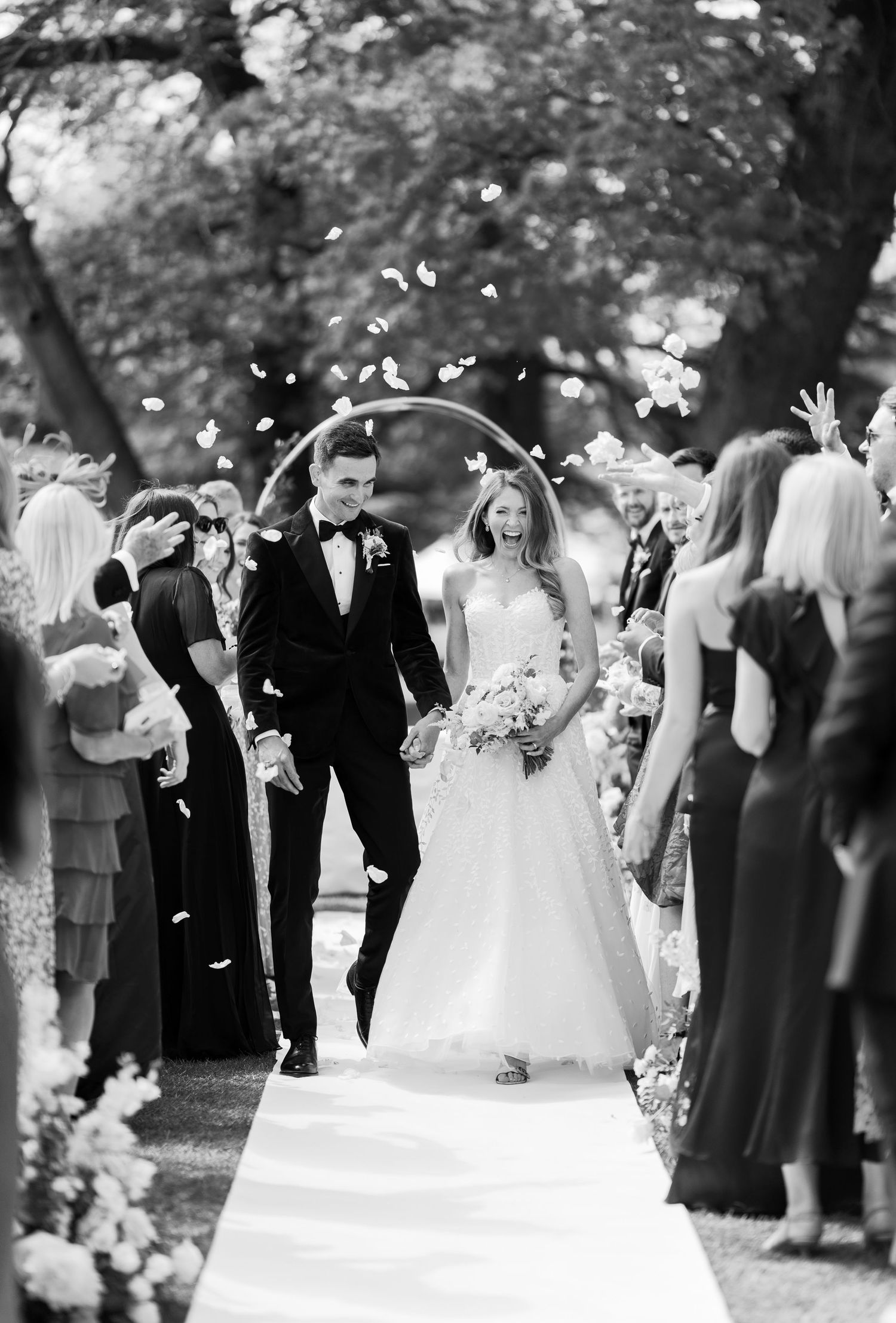 Newlyweds walk down aisle as guests toss flower petals during outdoor wedding recessional.