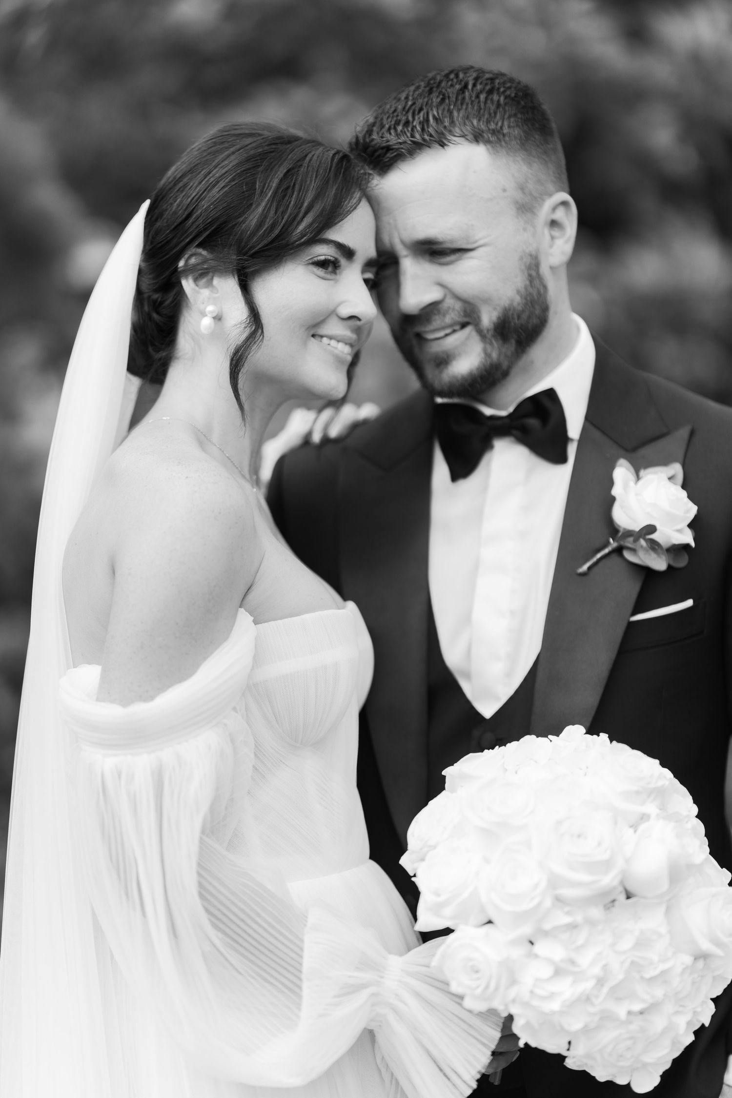 Black and white wedding photo of smiling bride in white gown and groom in tuxedo sharing an intimate moment.