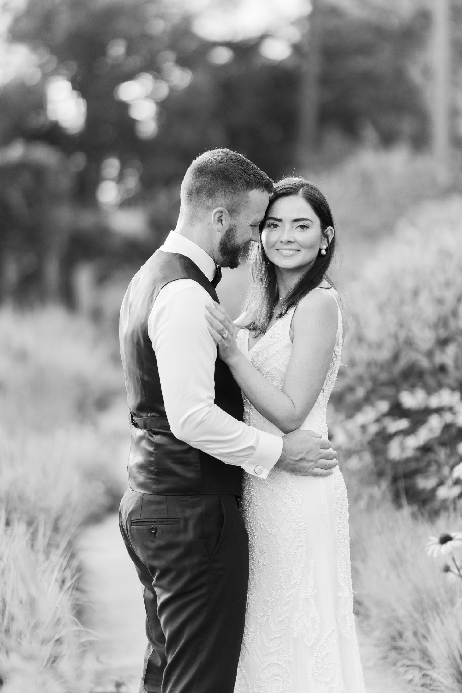 Black and white wedding photo of bride and groom embracing outdoors, bride smiling at camera in elegant white gown.