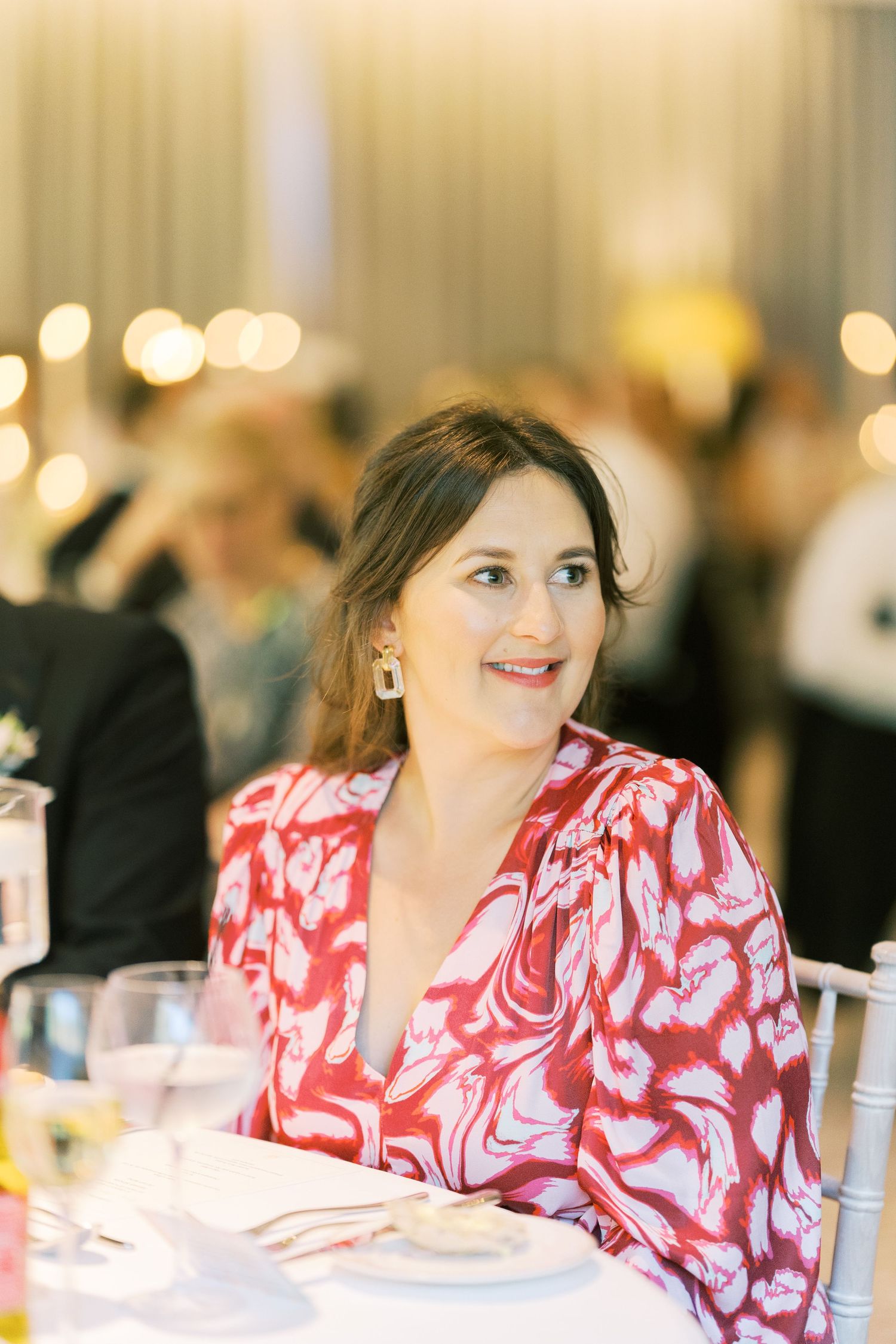 Woman in red floral dress smiling at elegant event dinner table with bokeh lights background.