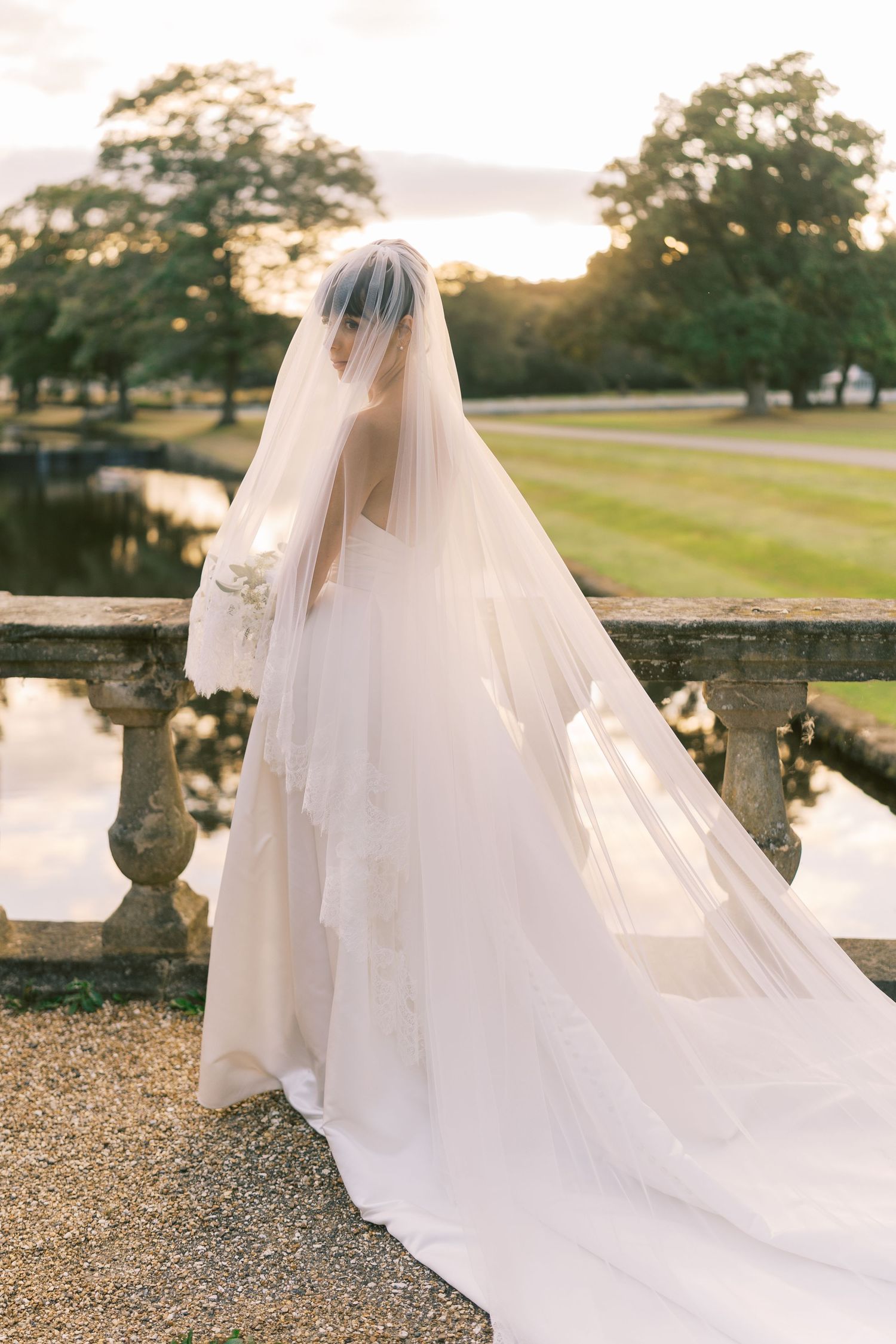 Bride in a flowing white gown and layered veil stands at a stone balustrade overlooking a serene garden at sunset.