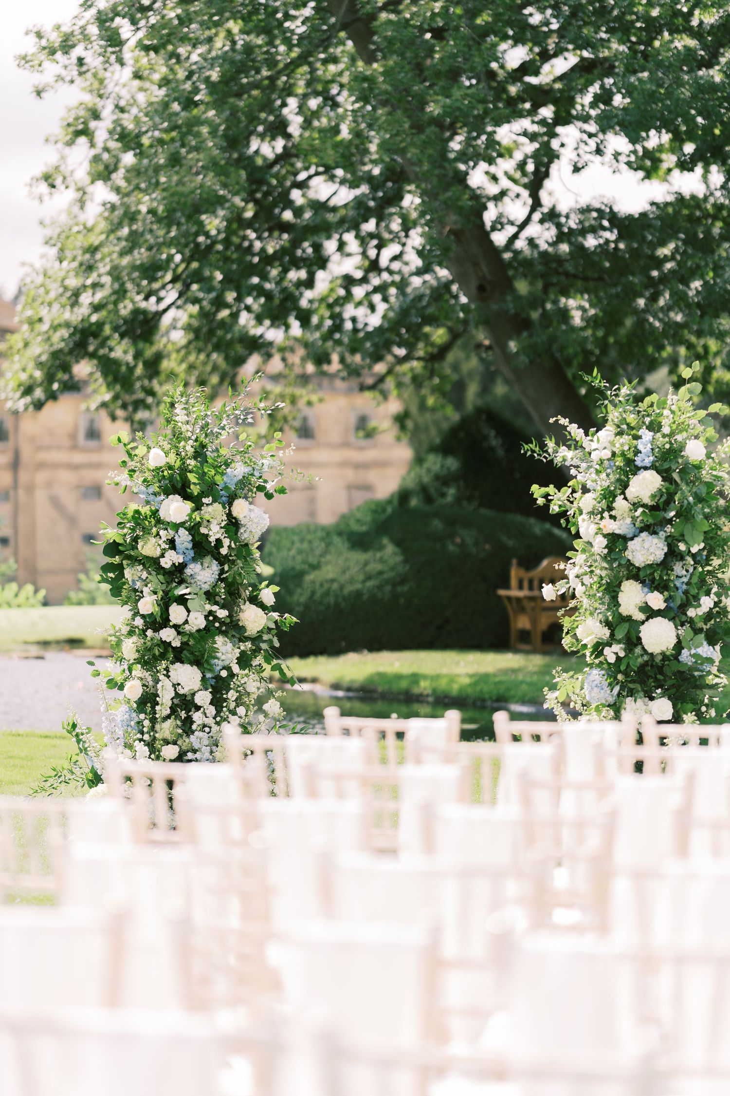 Elegant outdoor wedding ceremony setup with floral arch of white and blue blooms framing chiavari chairs on lawn.