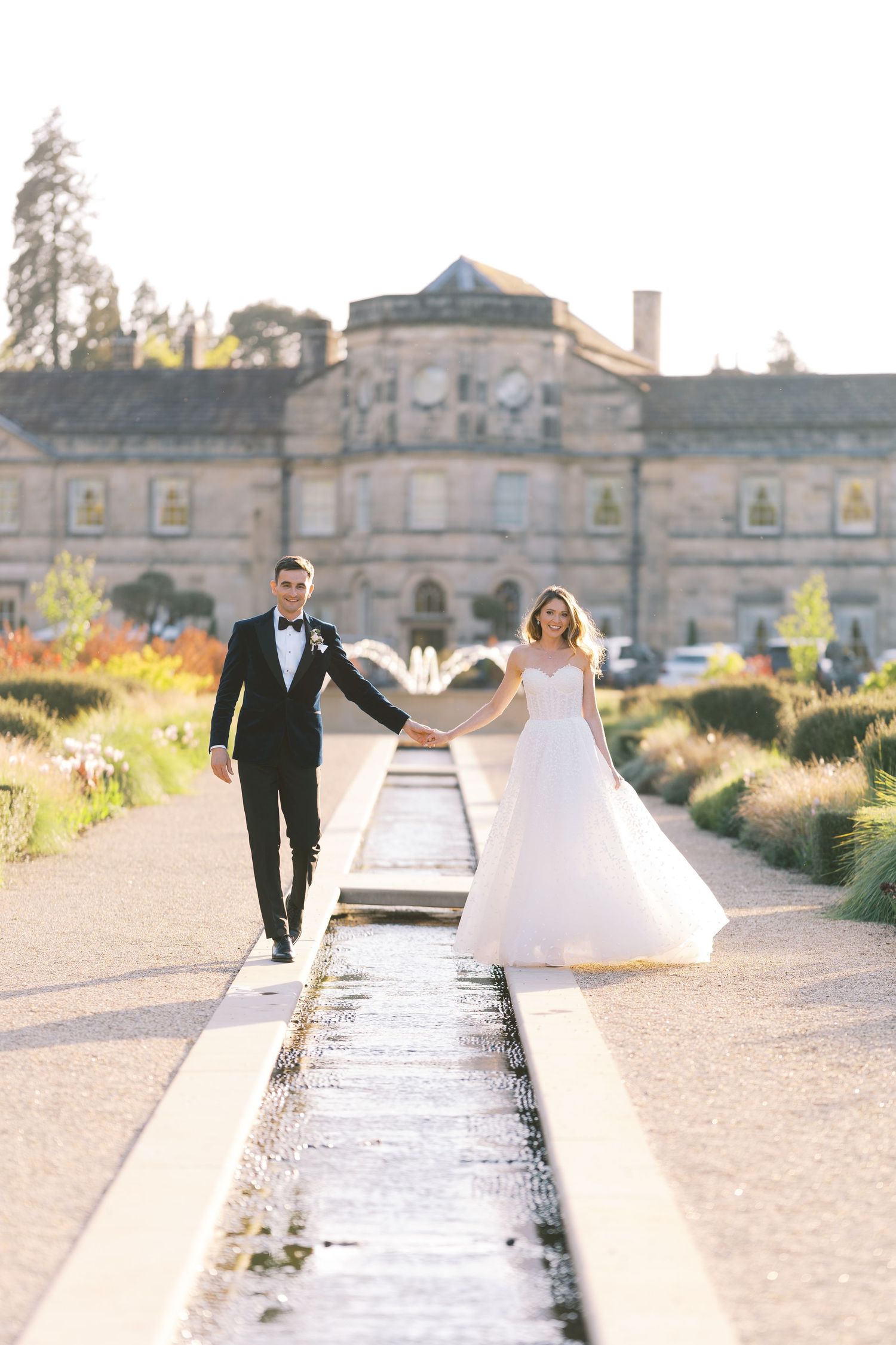 Bride and groom walk hand-in-hand along a water feature path at a grand estate wedding venue in golden hour light.