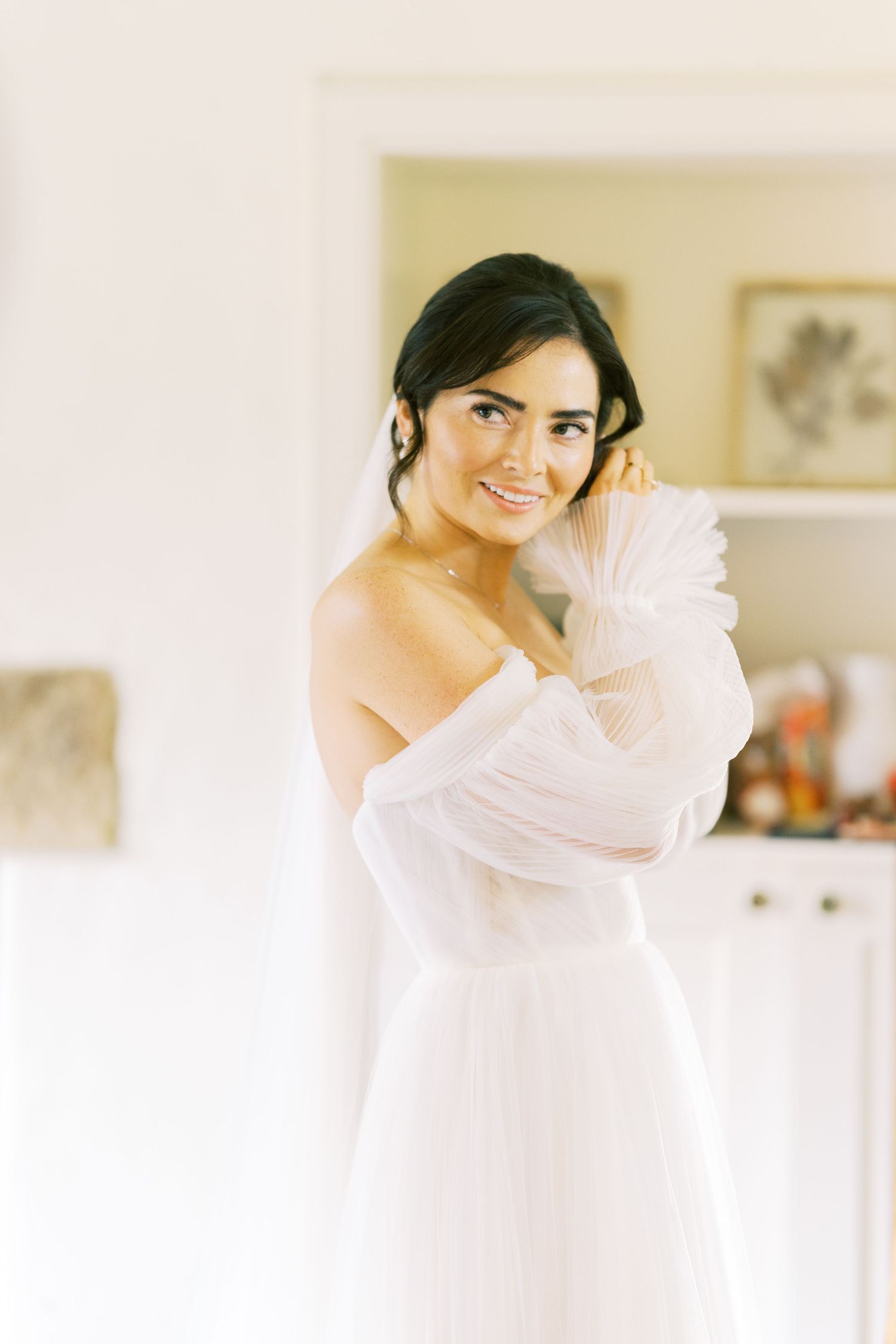 Elegant bride in off-shoulder white gown with sheer sleeves smiling softly in a bright, airy room.