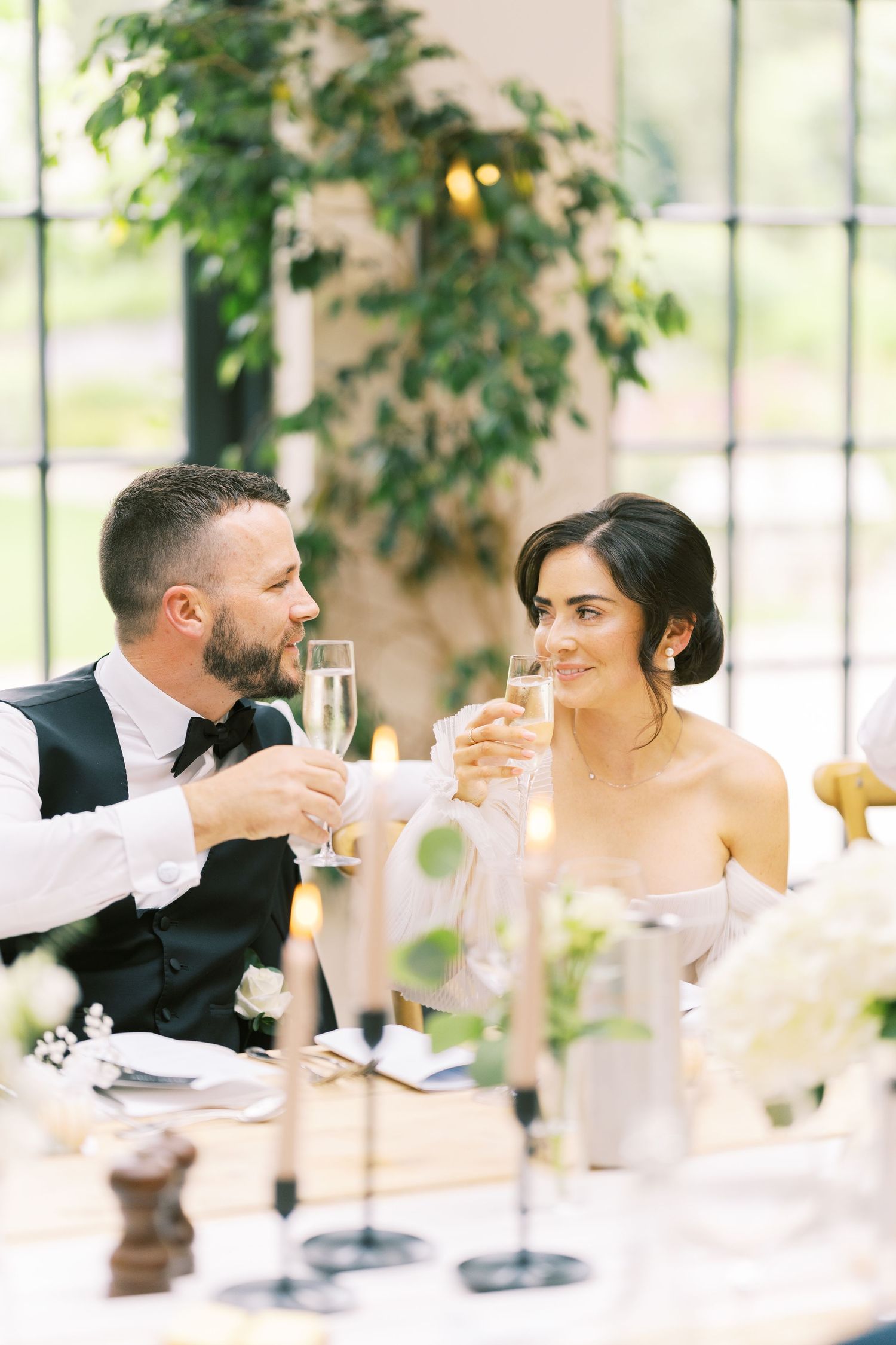 Bride and groom toast with champagne glasses at elegant wedding reception table with candles and greenery.