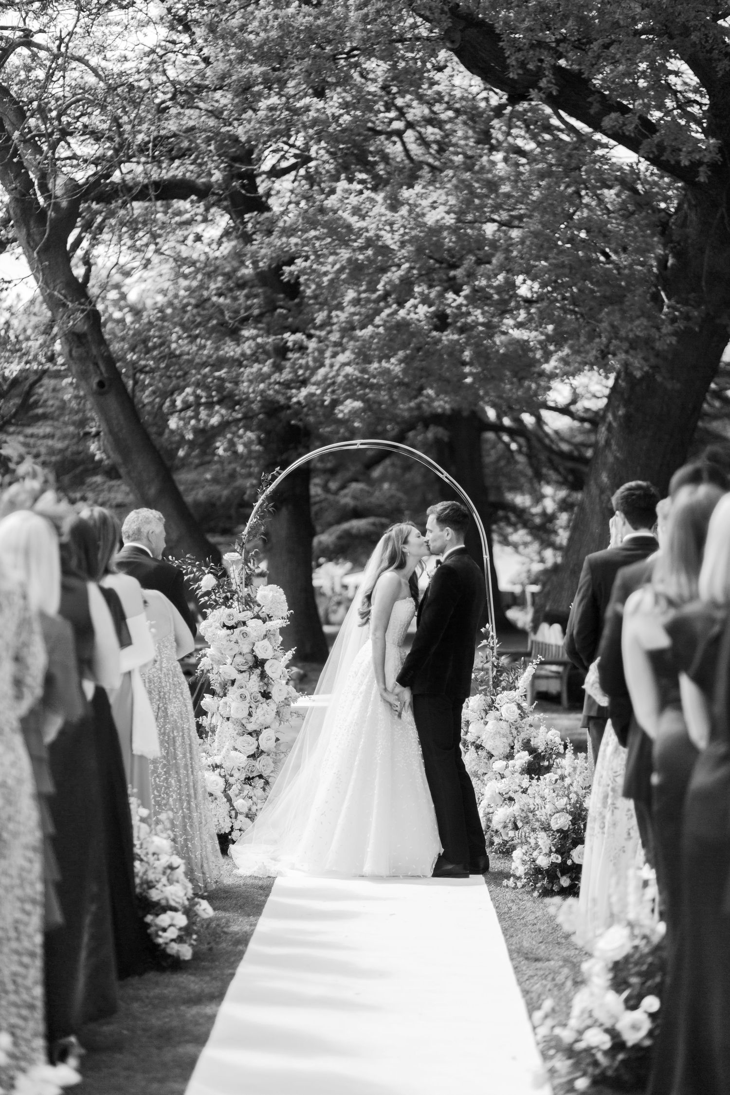 Bride and groom share first kiss under floral arch at elegant outdoor wedding ceremony in black and white.