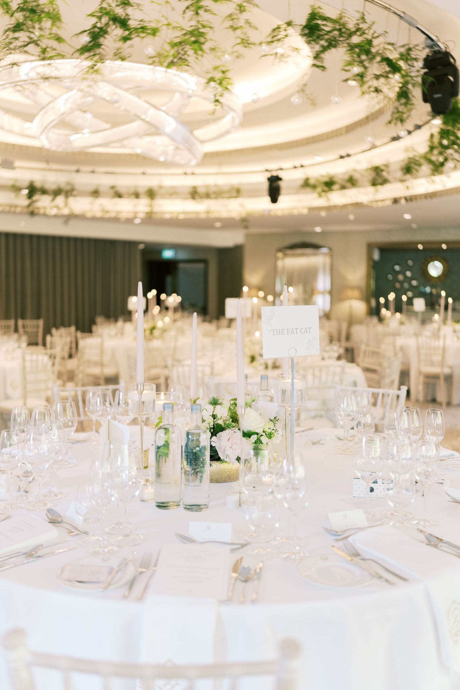 Luxurious wedding reception ballroom with white round tables, crystal glassware, candles, and greenery ceiling installation.