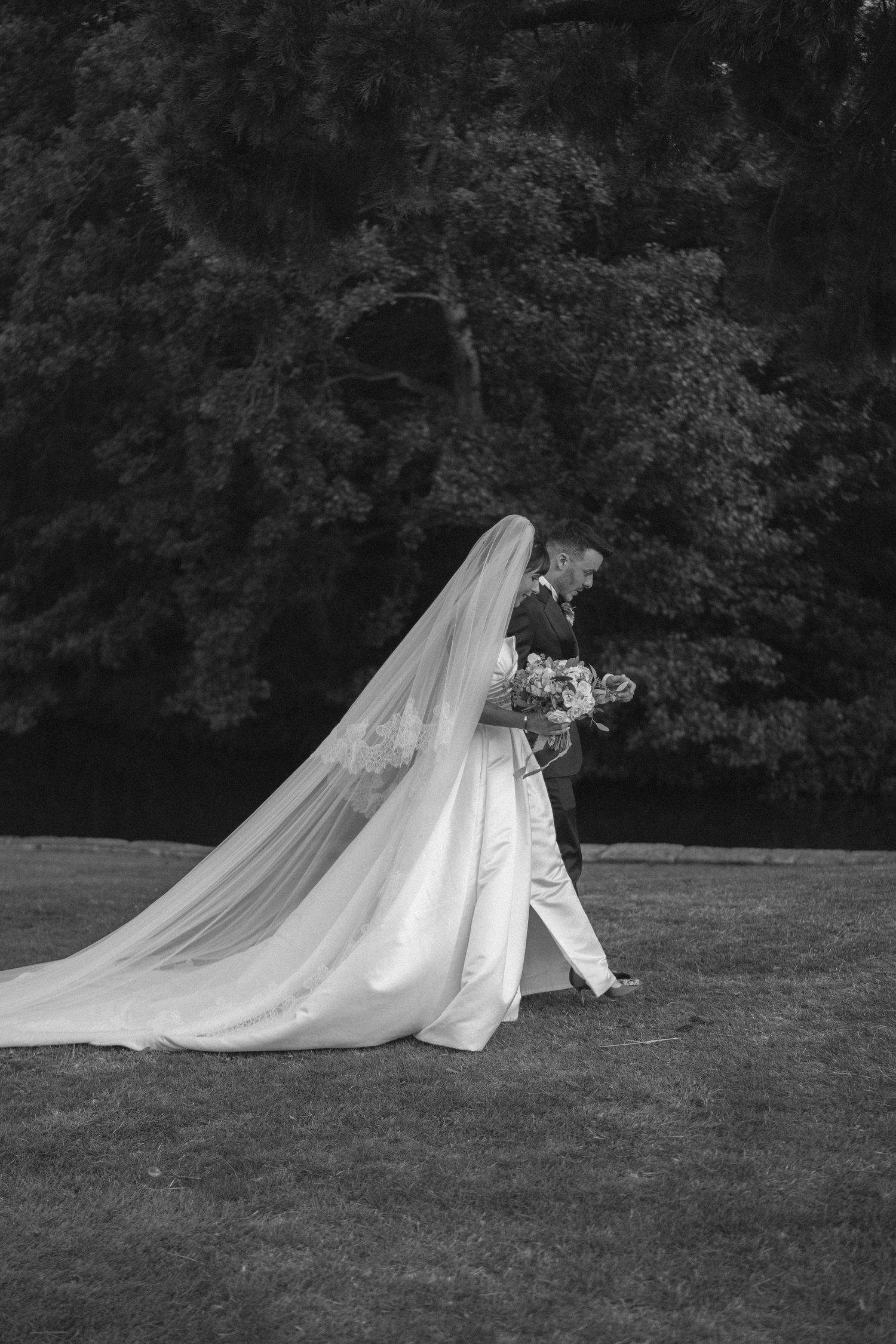 Black and white photo of a bride and groom walking on grass, her long veil trailing behind them.