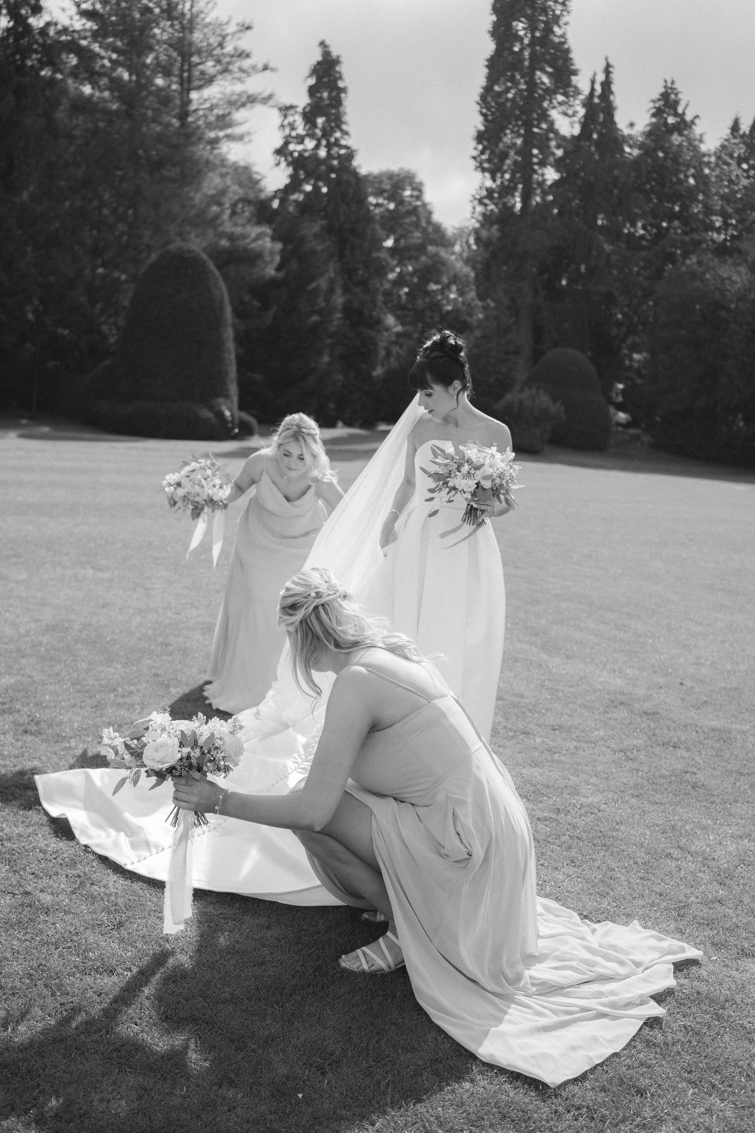 Black and white wedding photo of bridesmaids arranging bride's veil and train on garden lawn.
