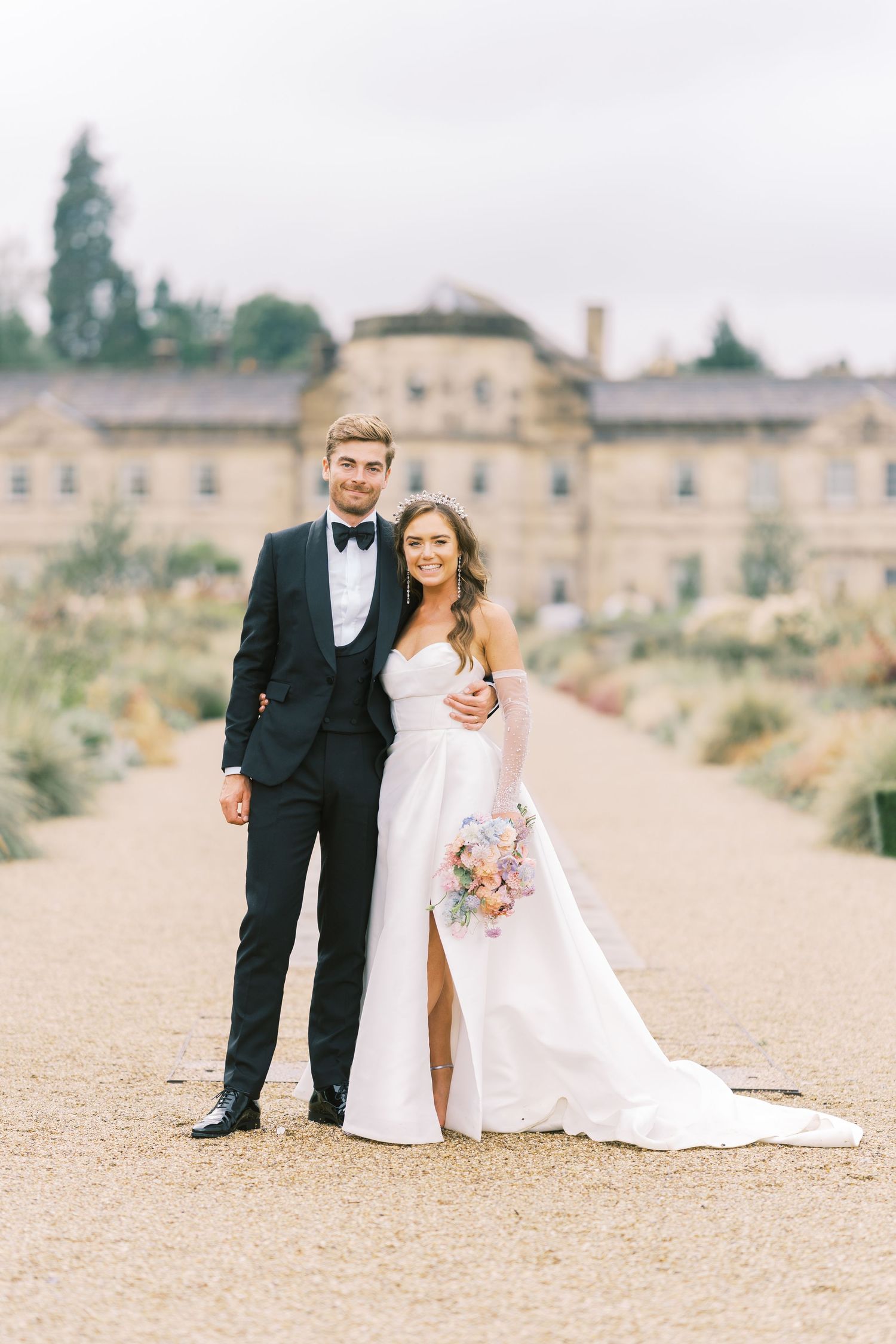 Elegant bride and groom posing together on a gravel path in front of a grand estate on their wedding day.