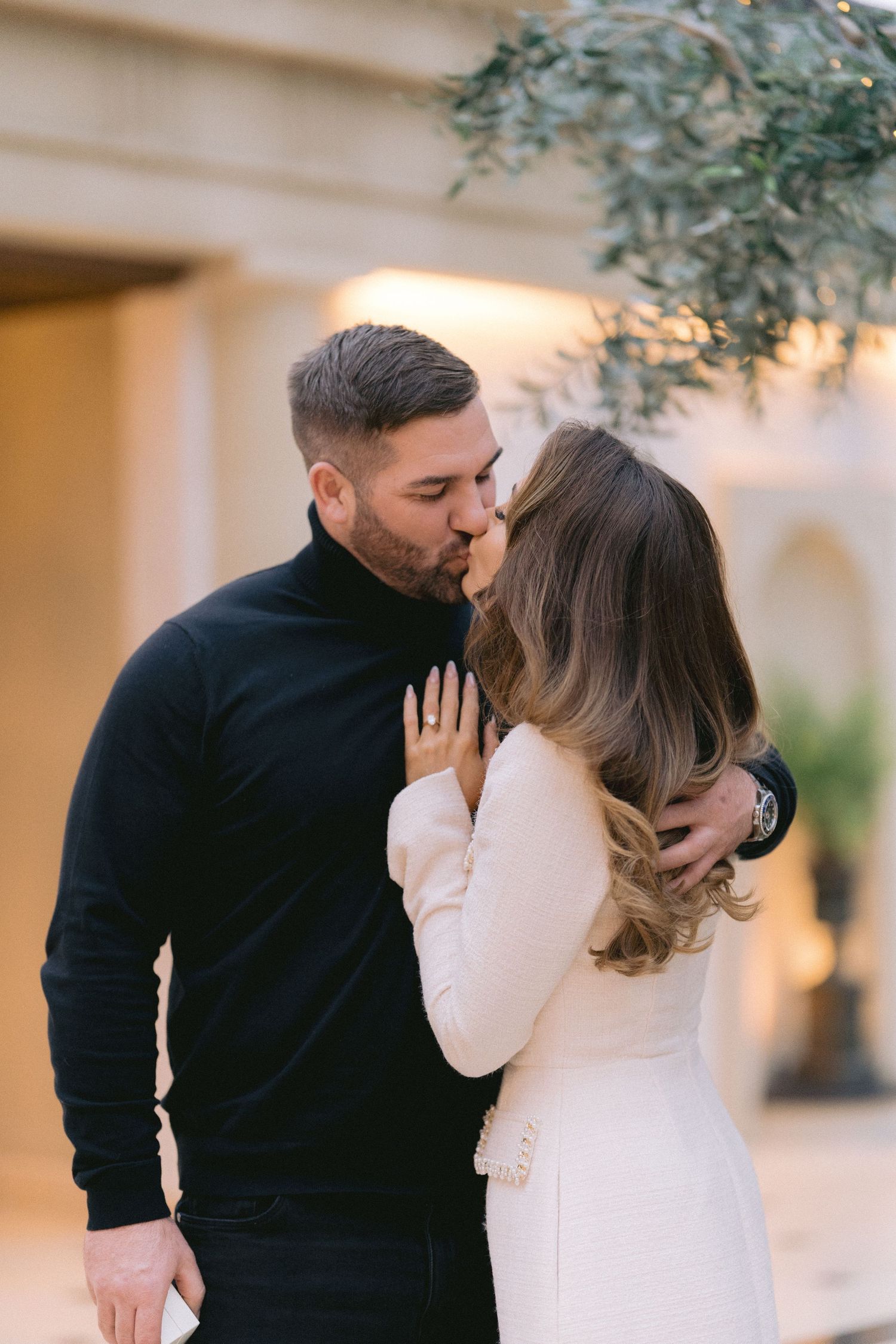 Newly engaged couple sharing a kiss outdoors near an olive tree, woman displaying engagement ring.