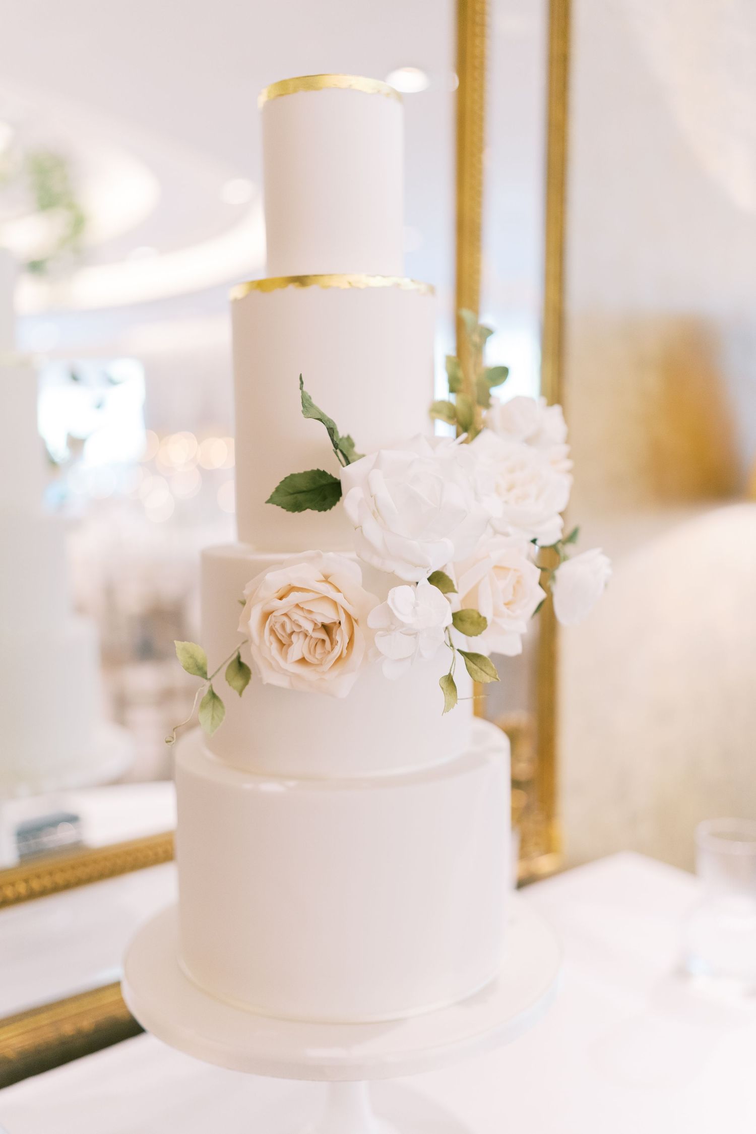 Four-tier white wedding cake with gold accents and fresh blush roses and white peony sugar flowers.