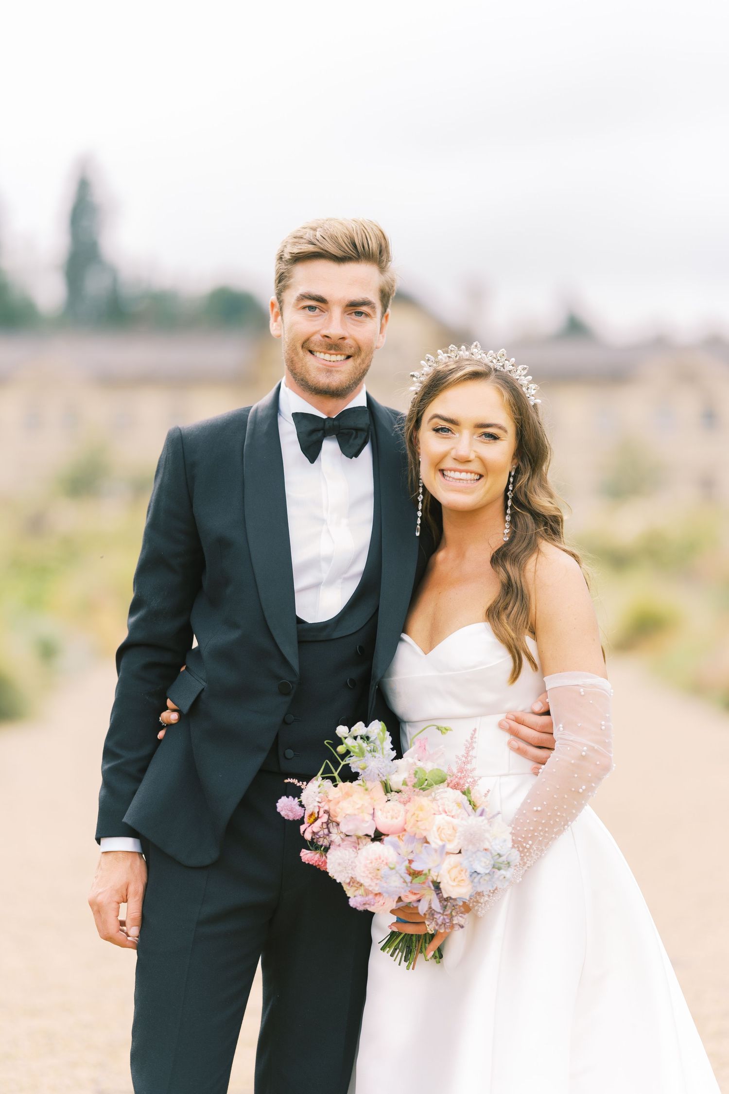 Smiling bride and groom posing outdoors, bride in strapless gown with tiara, groom in black tuxedo with bow tie.