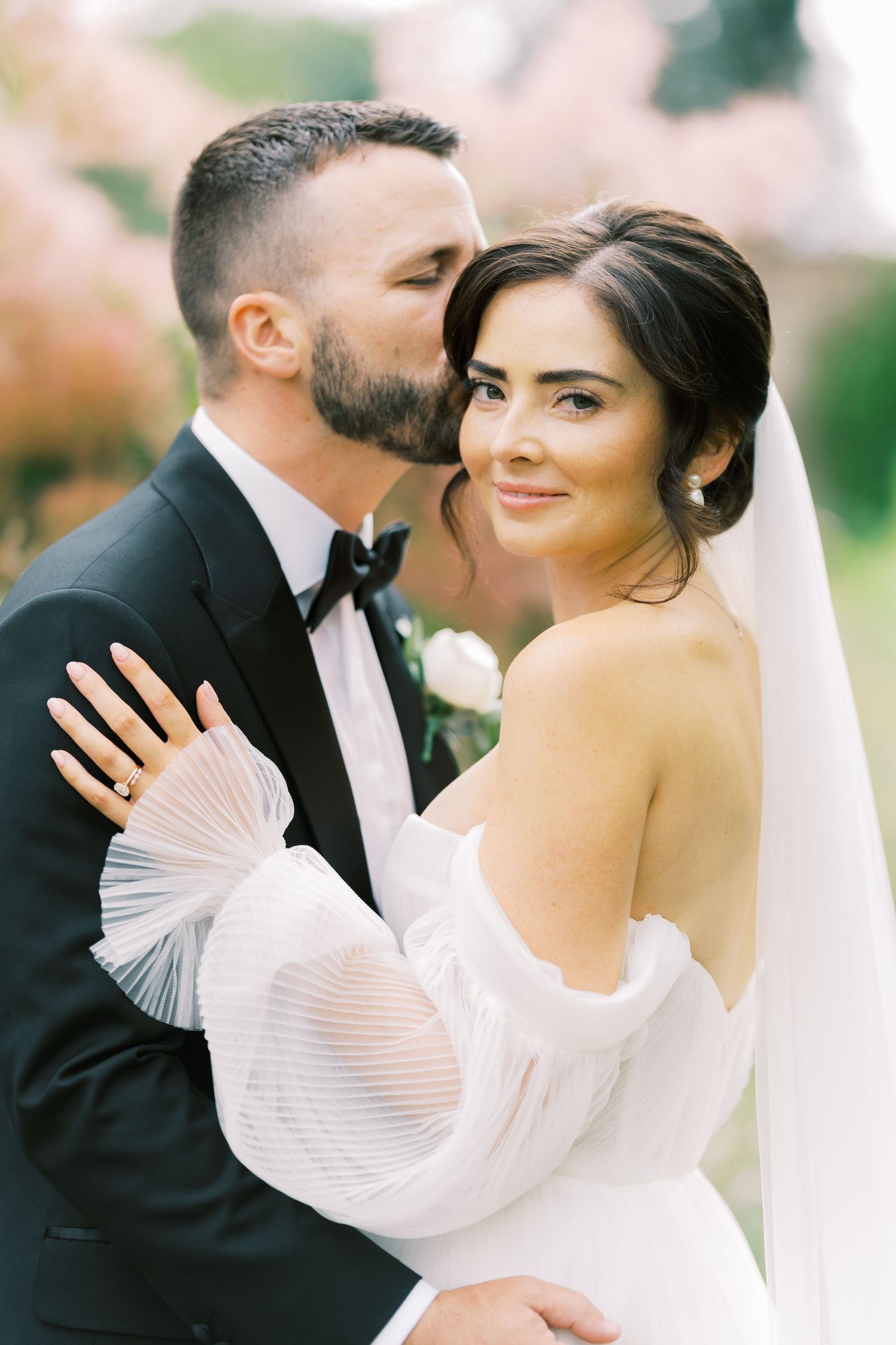 Groom in black tuxedo kissing bride's cheek, bride wearing off-shoulder white gown with veil, pink floral backdrop.