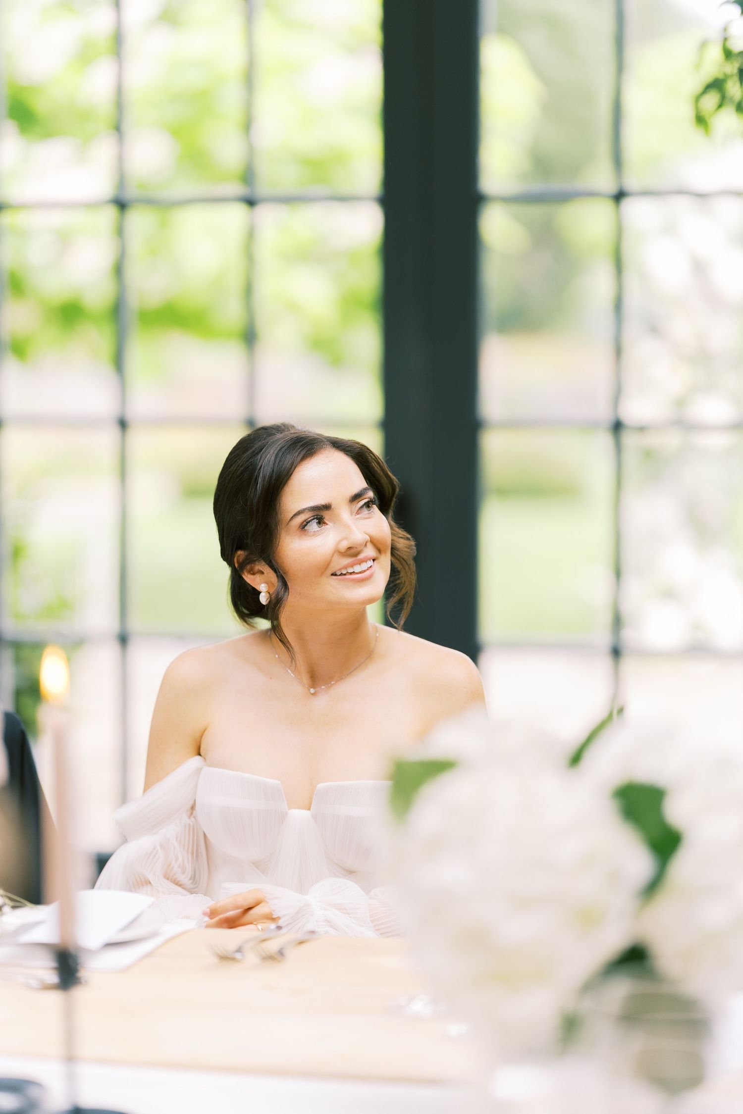 Elegant bride in off-shoulder white gown smiling at wedding reception table with floral arrangements.