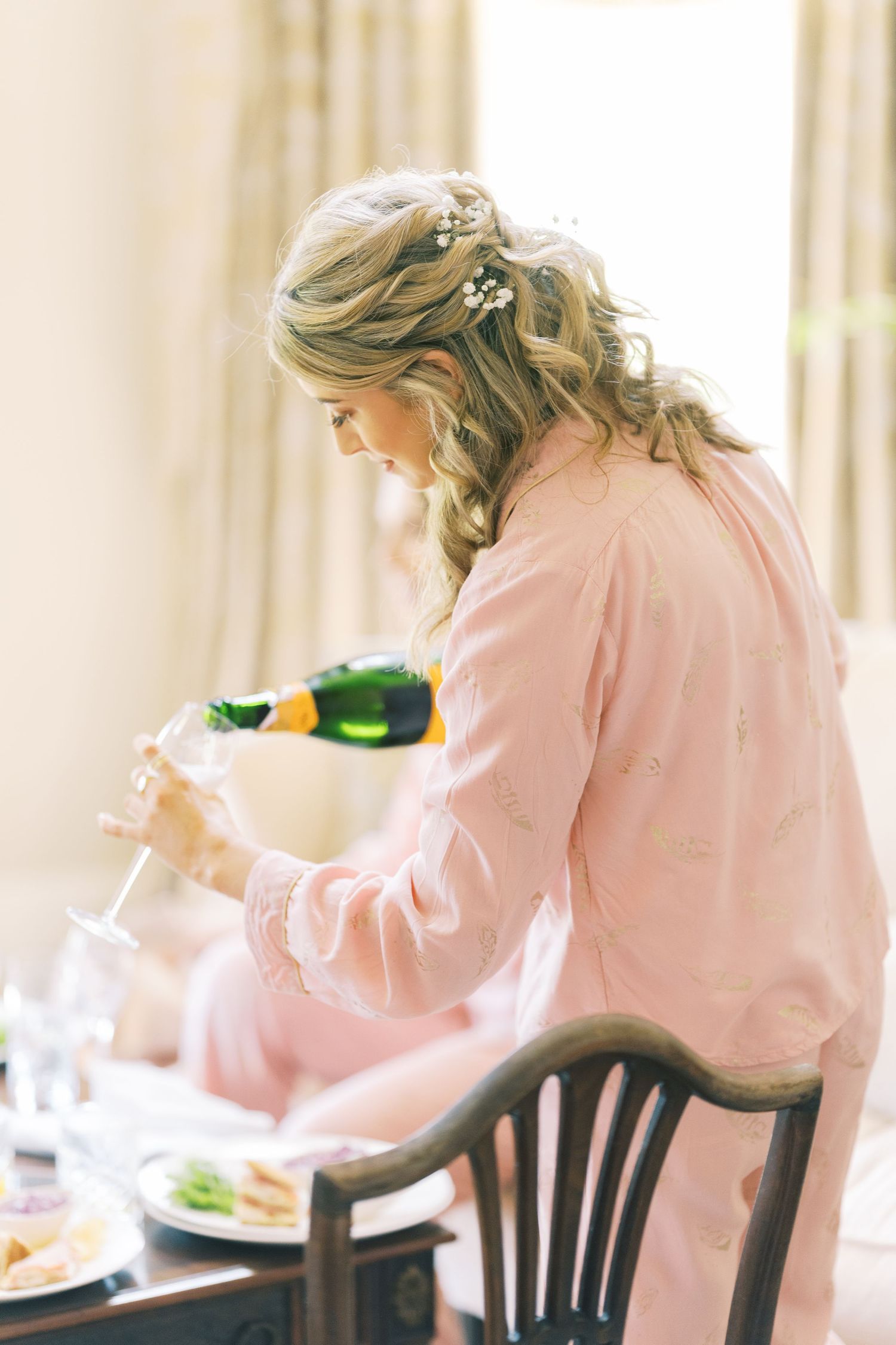 Bride in pink robe pouring champagne into a glass while getting ready on her wedding day.