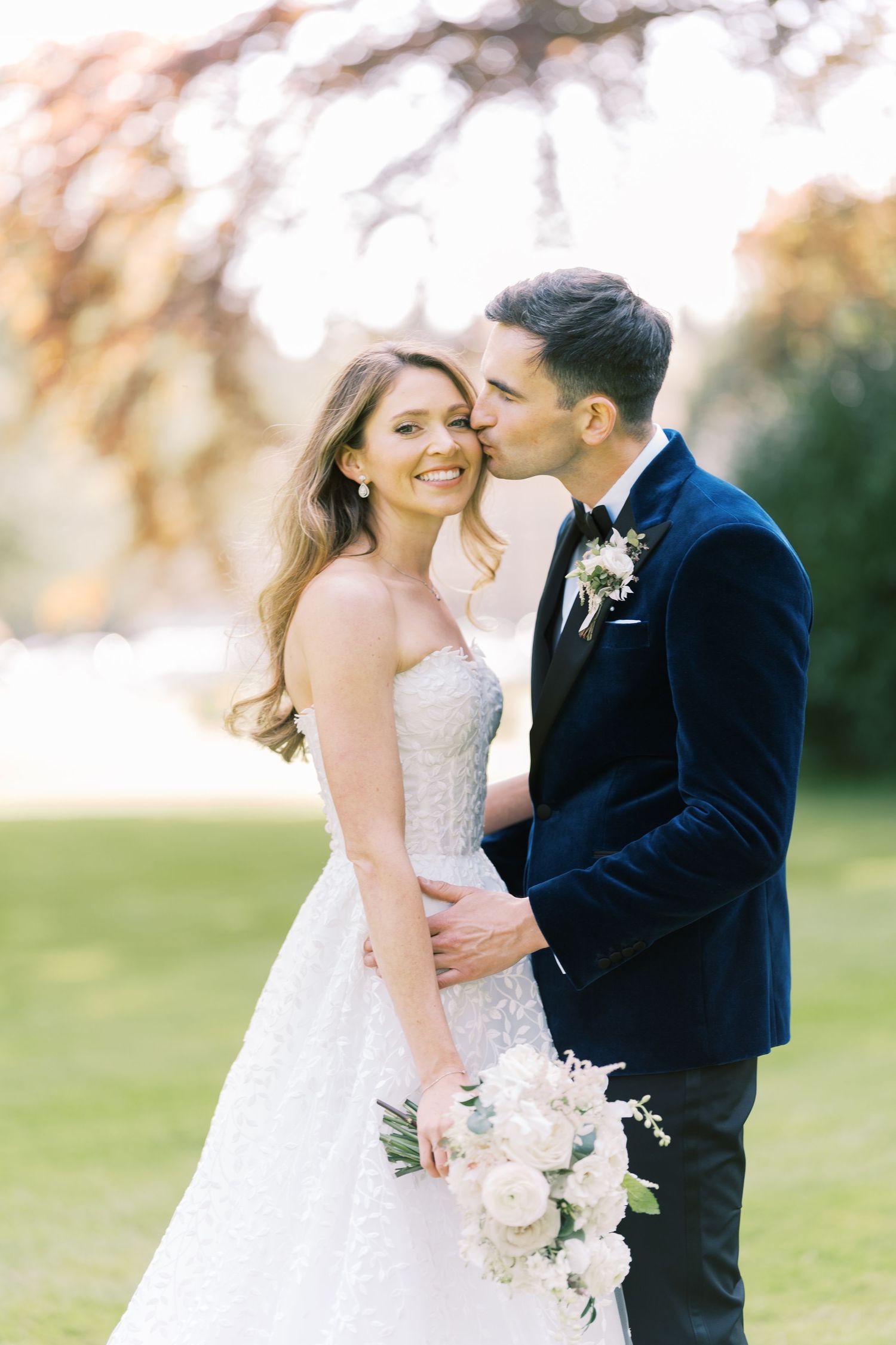Groom in navy tuxedo kisses bride in strapless beaded gown holding white bouquet outdoors at elegant wedding.