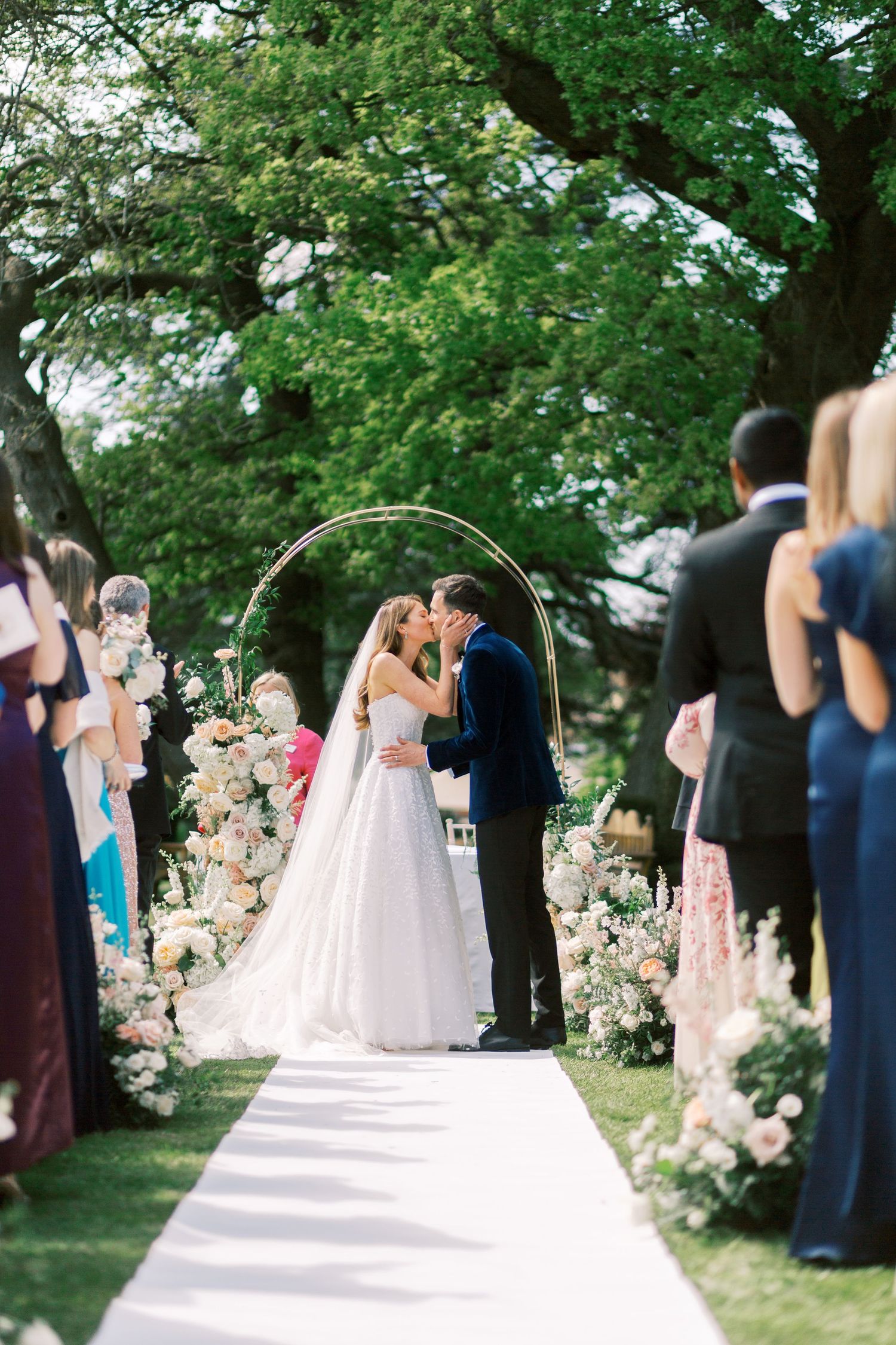 Bride and groom share first kiss under gold circular arch adorned with blush floral arrangements outdoors.
