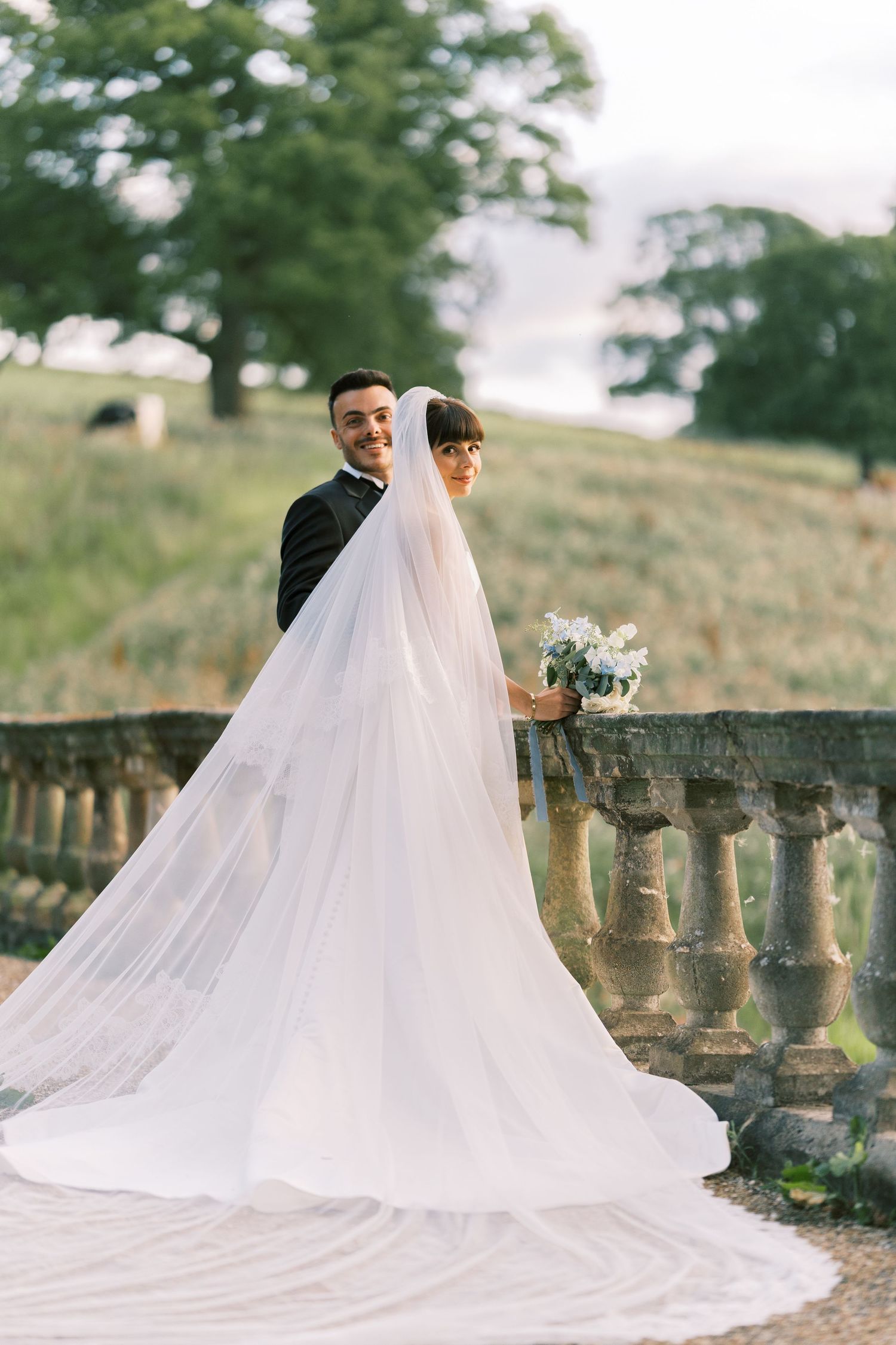 Smiling groom in tuxedo embraces bride in flowing white veil near stone balustrade outdoors.