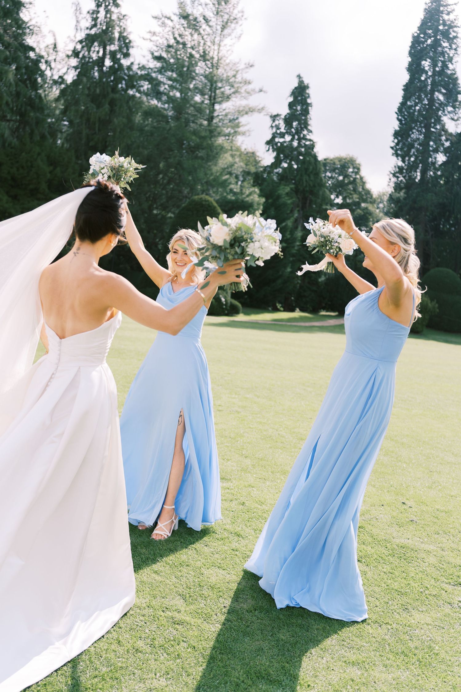 Bride in white gown with veil and two bridesmaids in light blue dresses celebrating outdoors on green lawn.