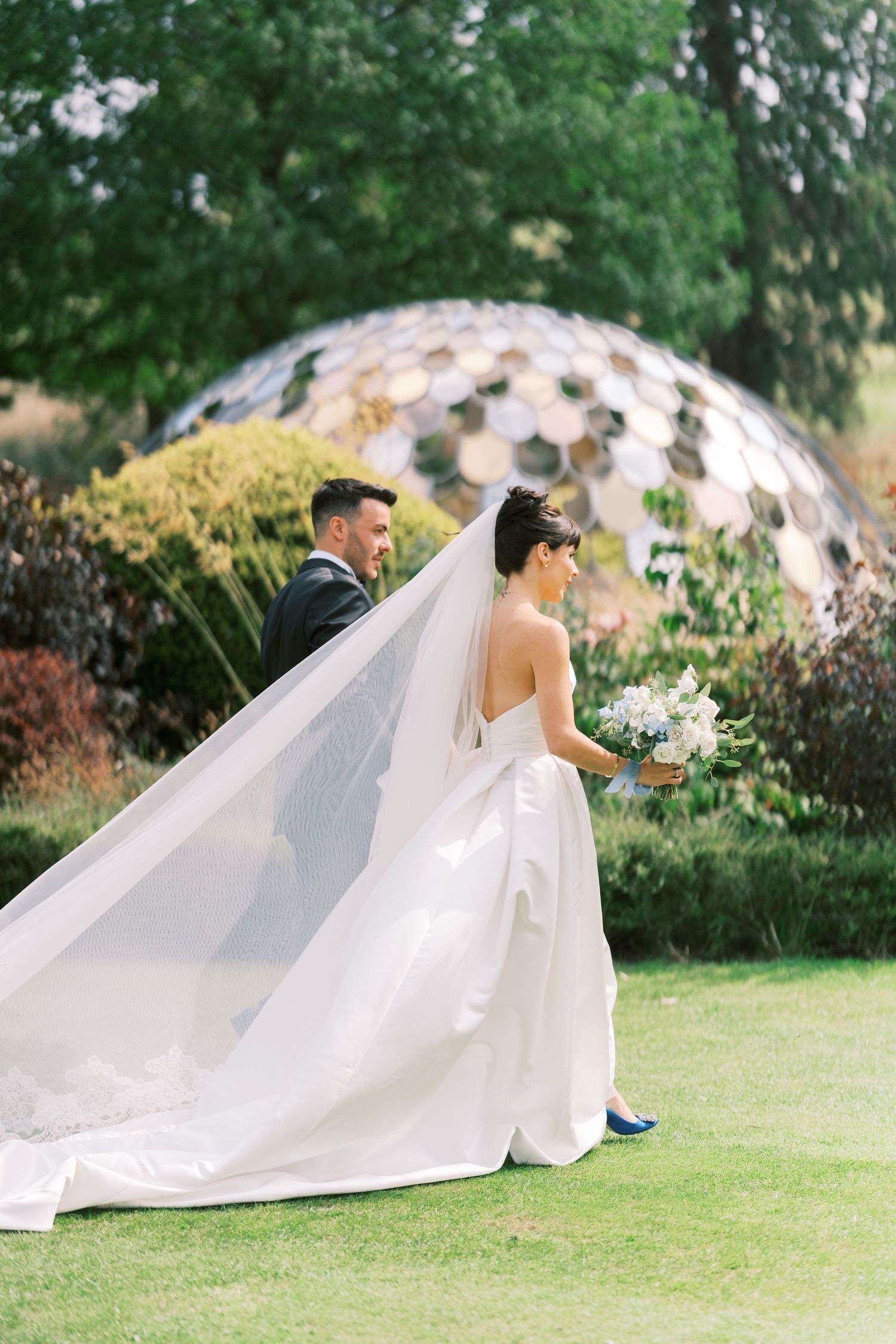 Bride in strapless gown with flowing veil and groom admire each other in sunlit garden with mirrored dome.
