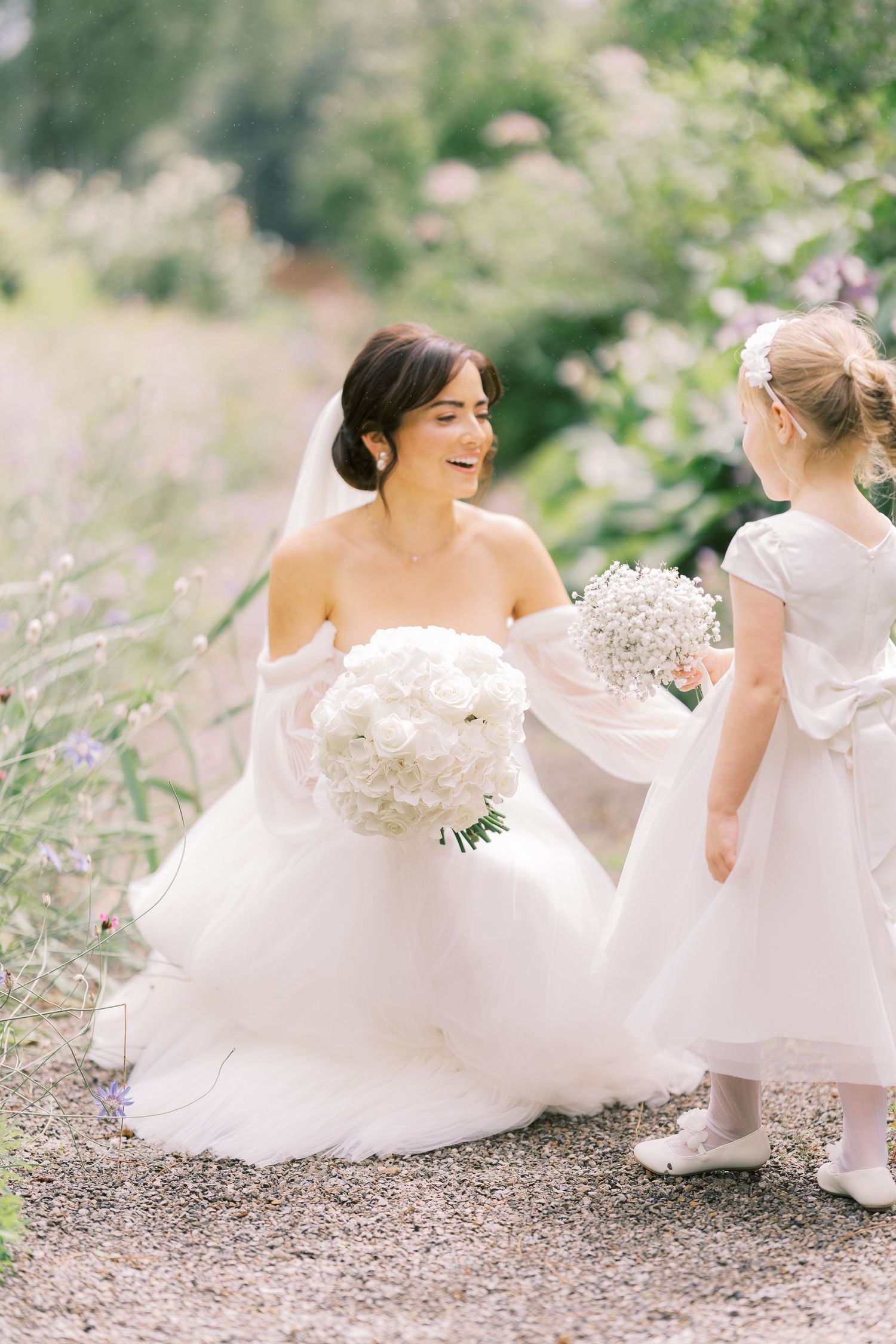 Bride in white tulle gown crouches to interact with young flower girl in garden setting.