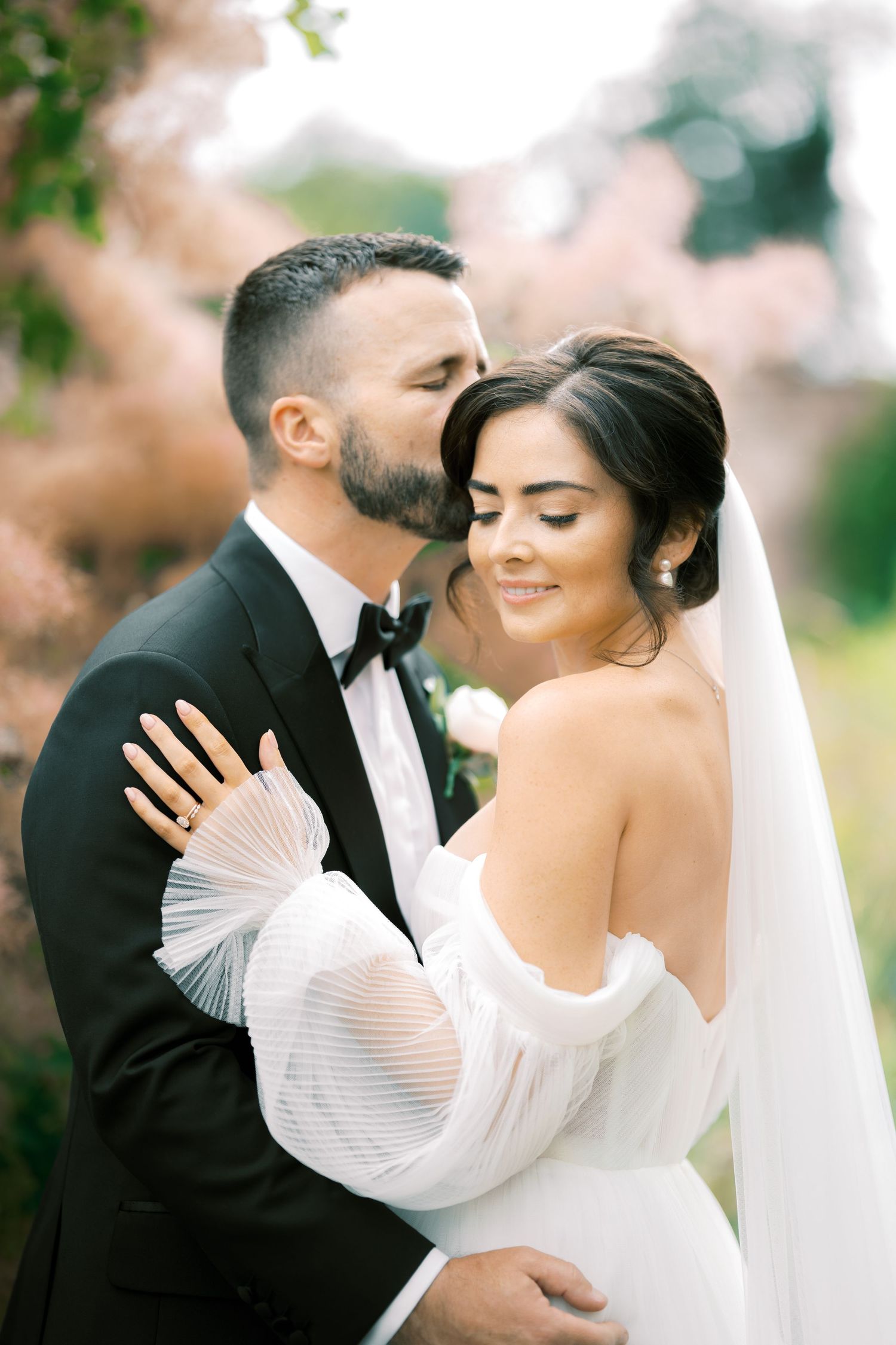 Groom in black tuxedo tenderly kisses bride in off-shoulder white gown with veil outdoors among pink blooms.