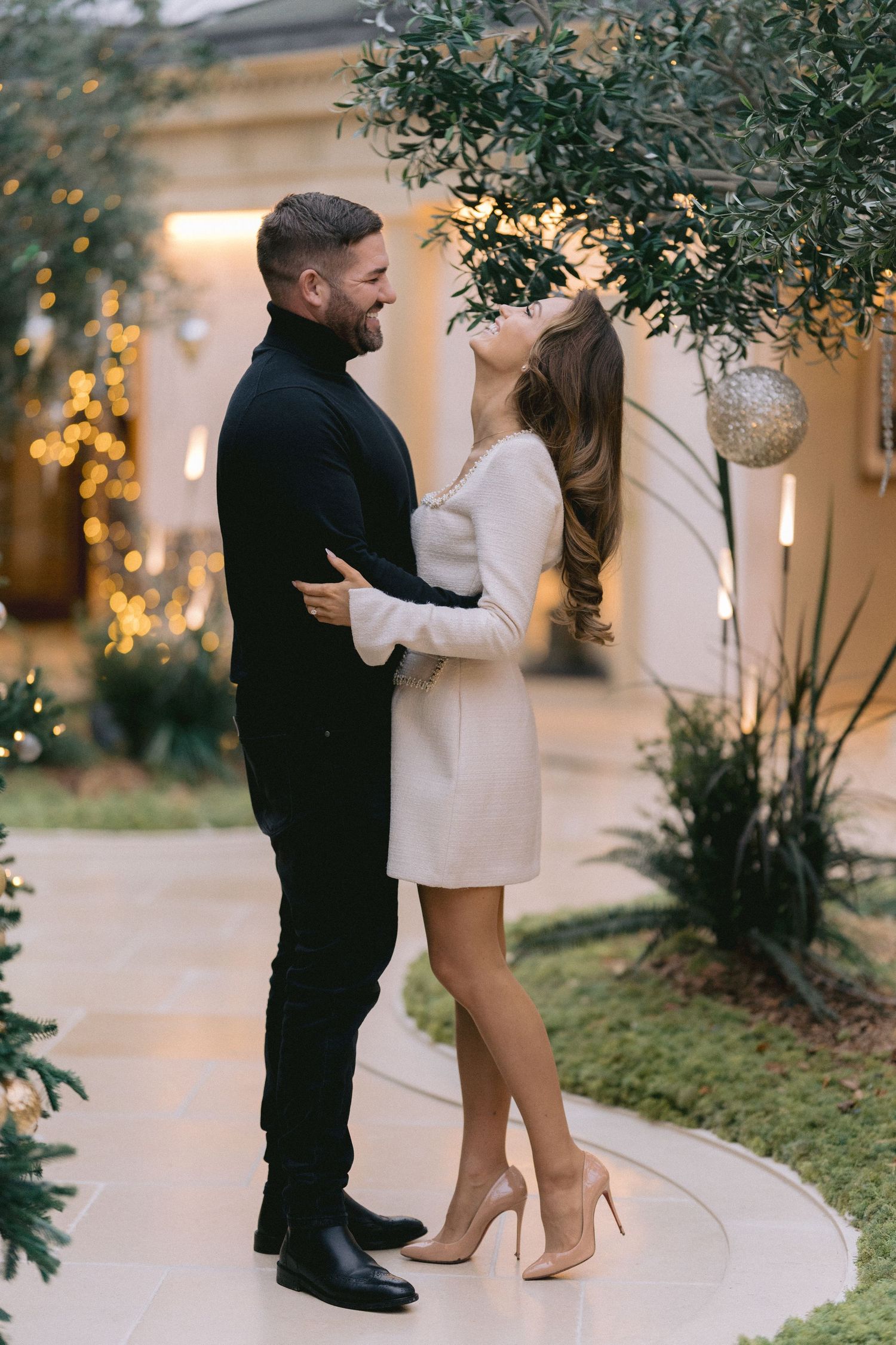 Couple sharing a joyful moment outdoors, smiling at each other near olive tree with warm bokeh lights.