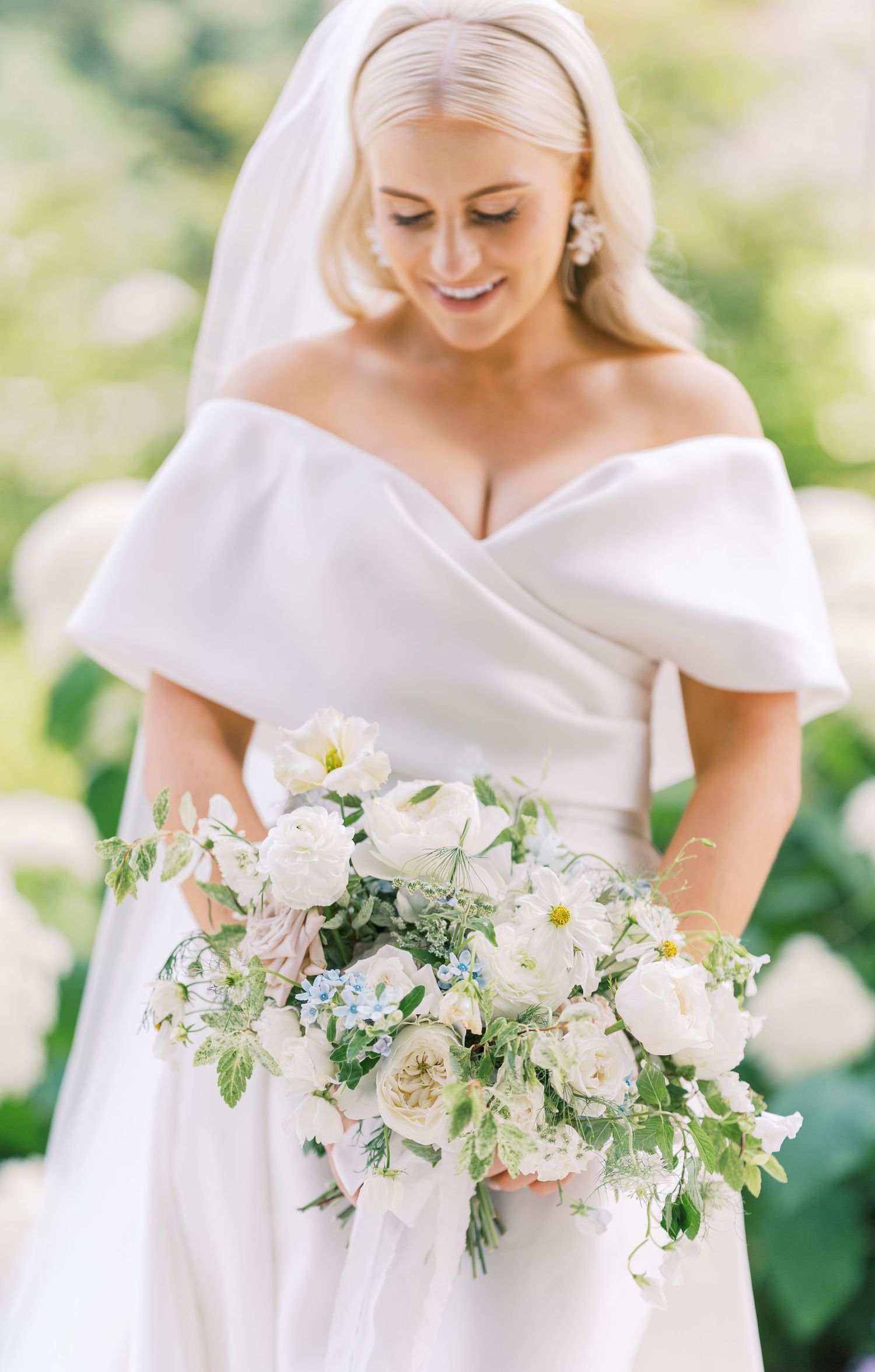 Radiant bride in off-shoulder gown smiles while holding elegant white and blush floral bouquet.