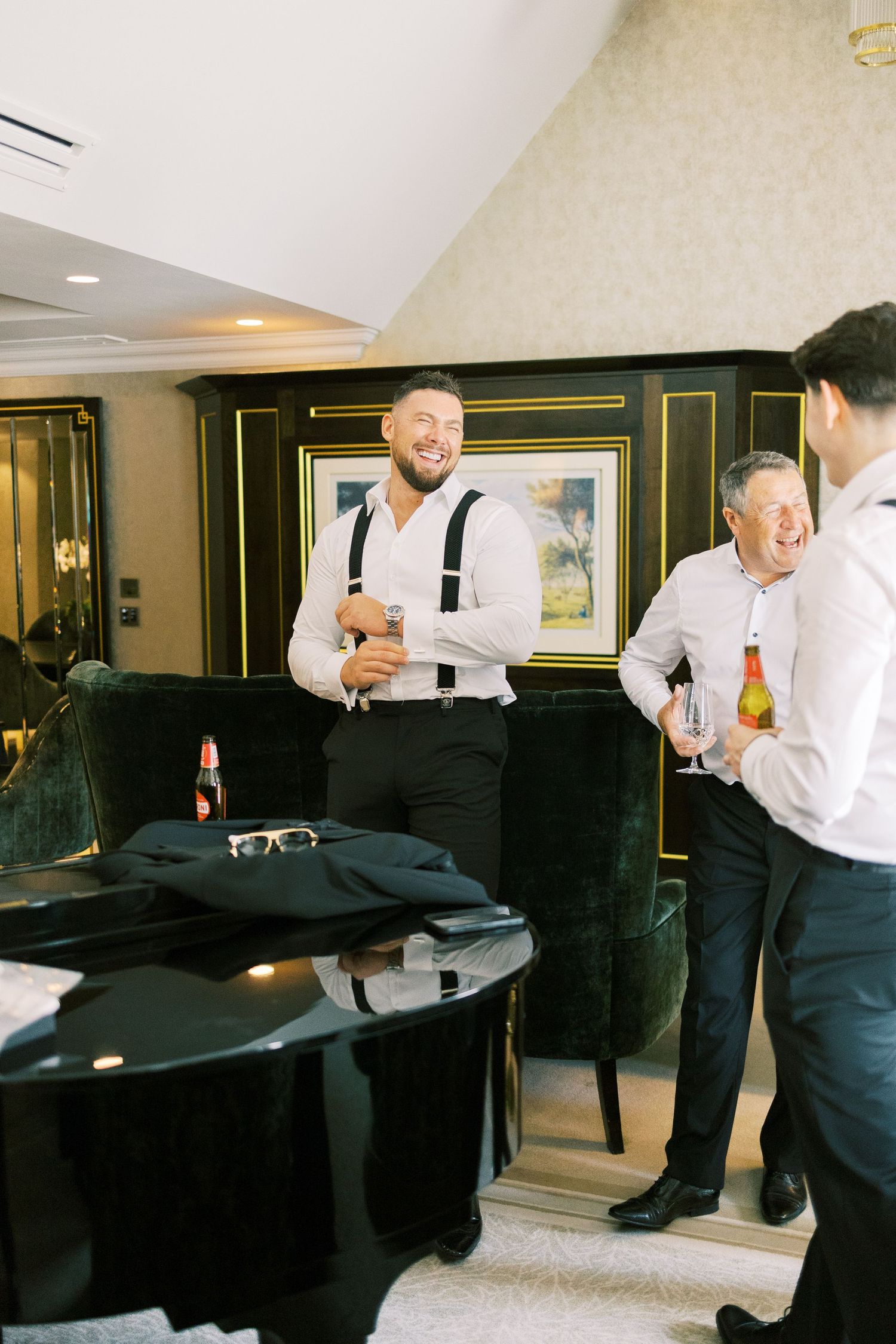 Groomsmen laughing and celebrating with drinks in an elegant hotel suite before a wedding.
