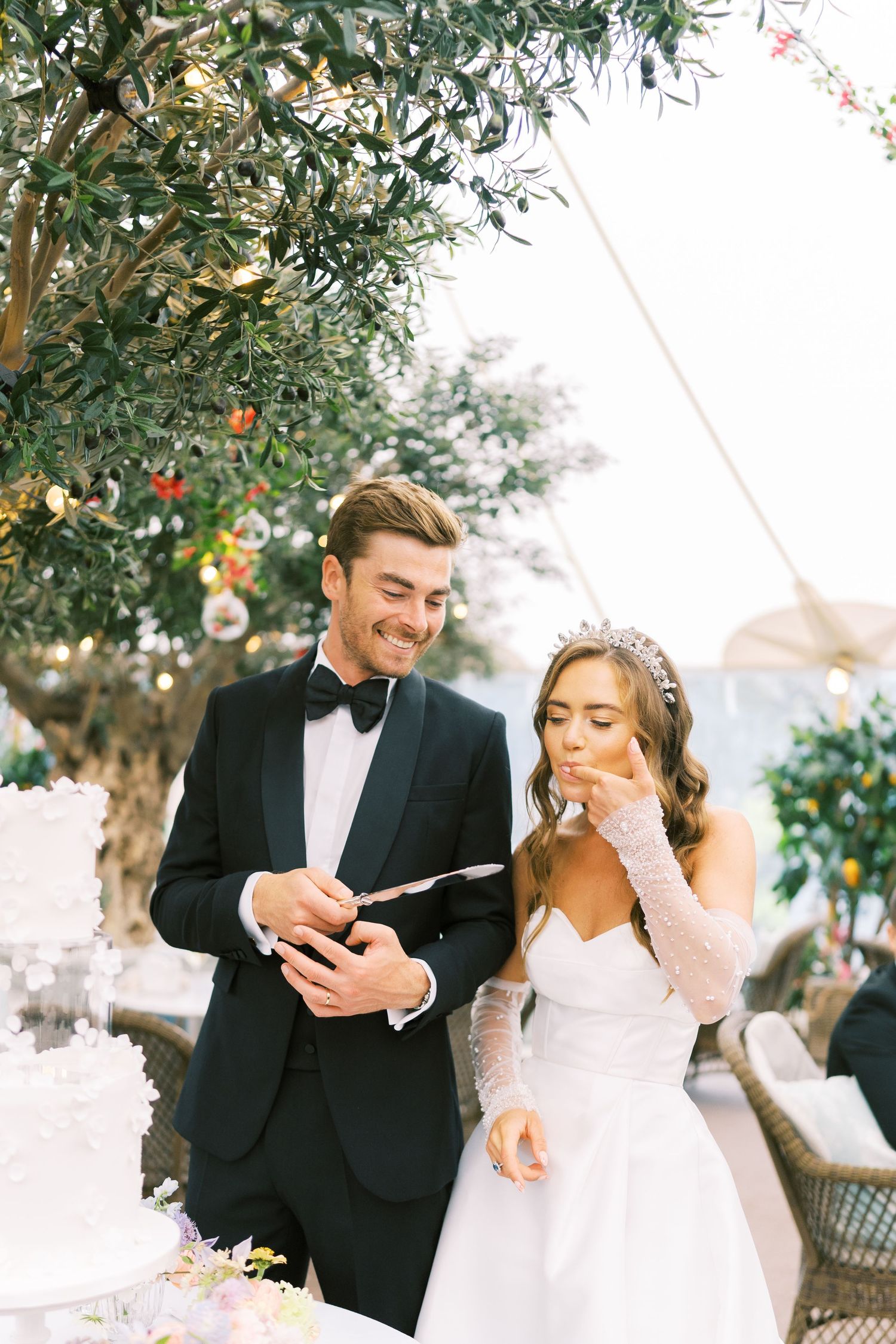Bride and groom cutting wedding cake outdoors under olive tree, bride tasting frosting in elegant white gown and tiara.