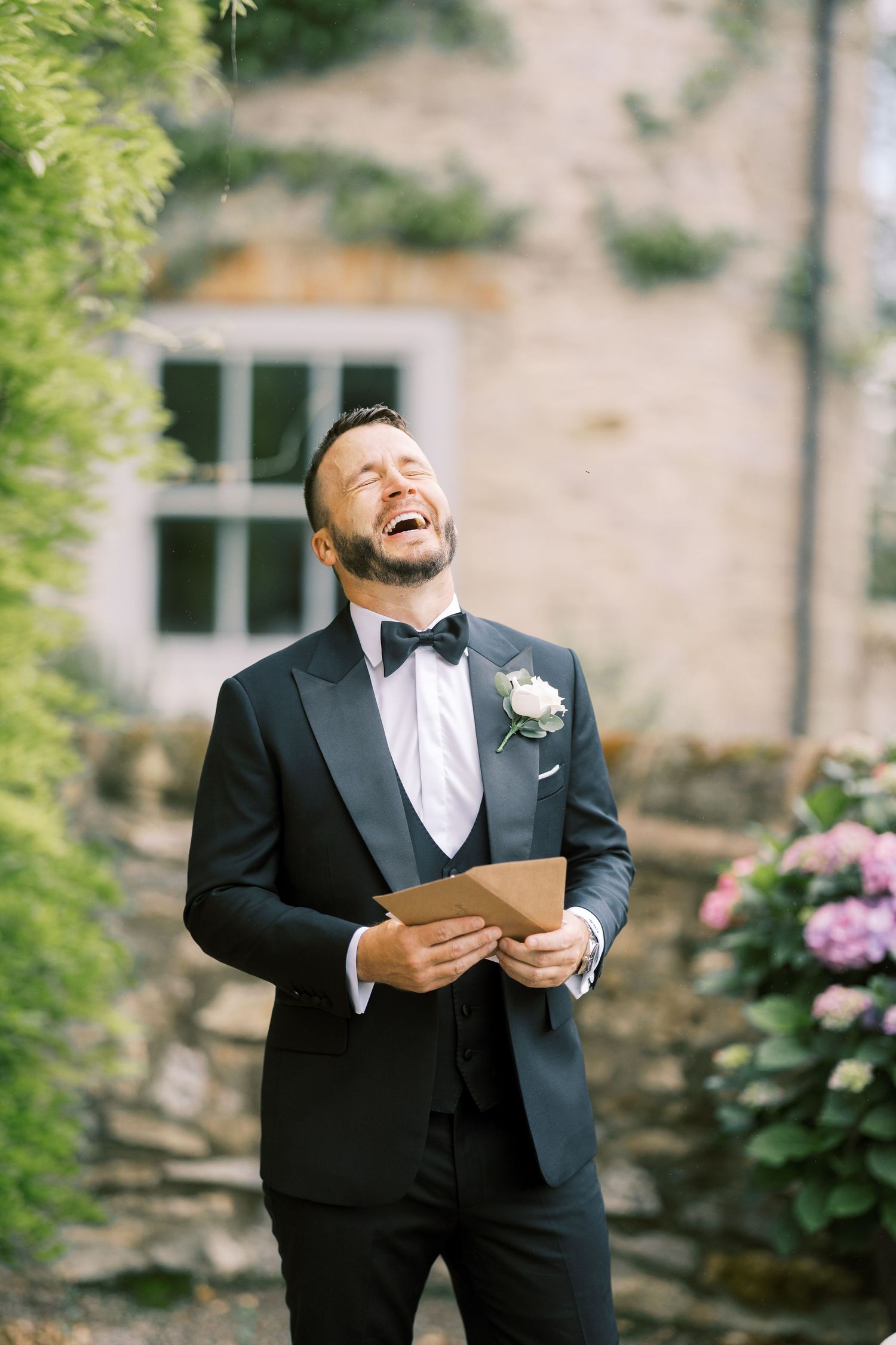 Groom in black tuxedo laughing joyfully while holding a card outdoors near stone building with flowers.
