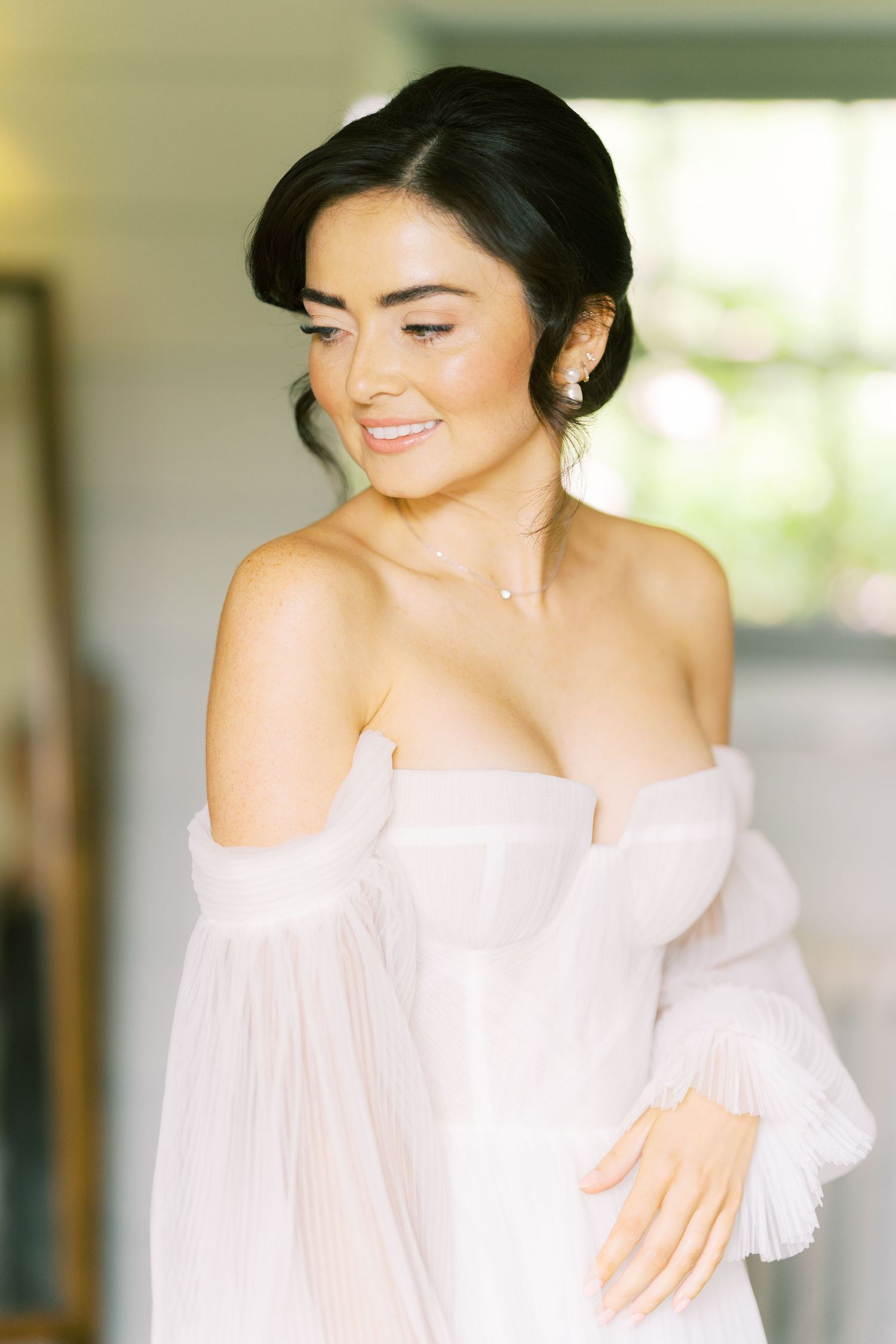 Beautiful bride in off-shoulder white gown with flowing sleeves smiling softly indoors.