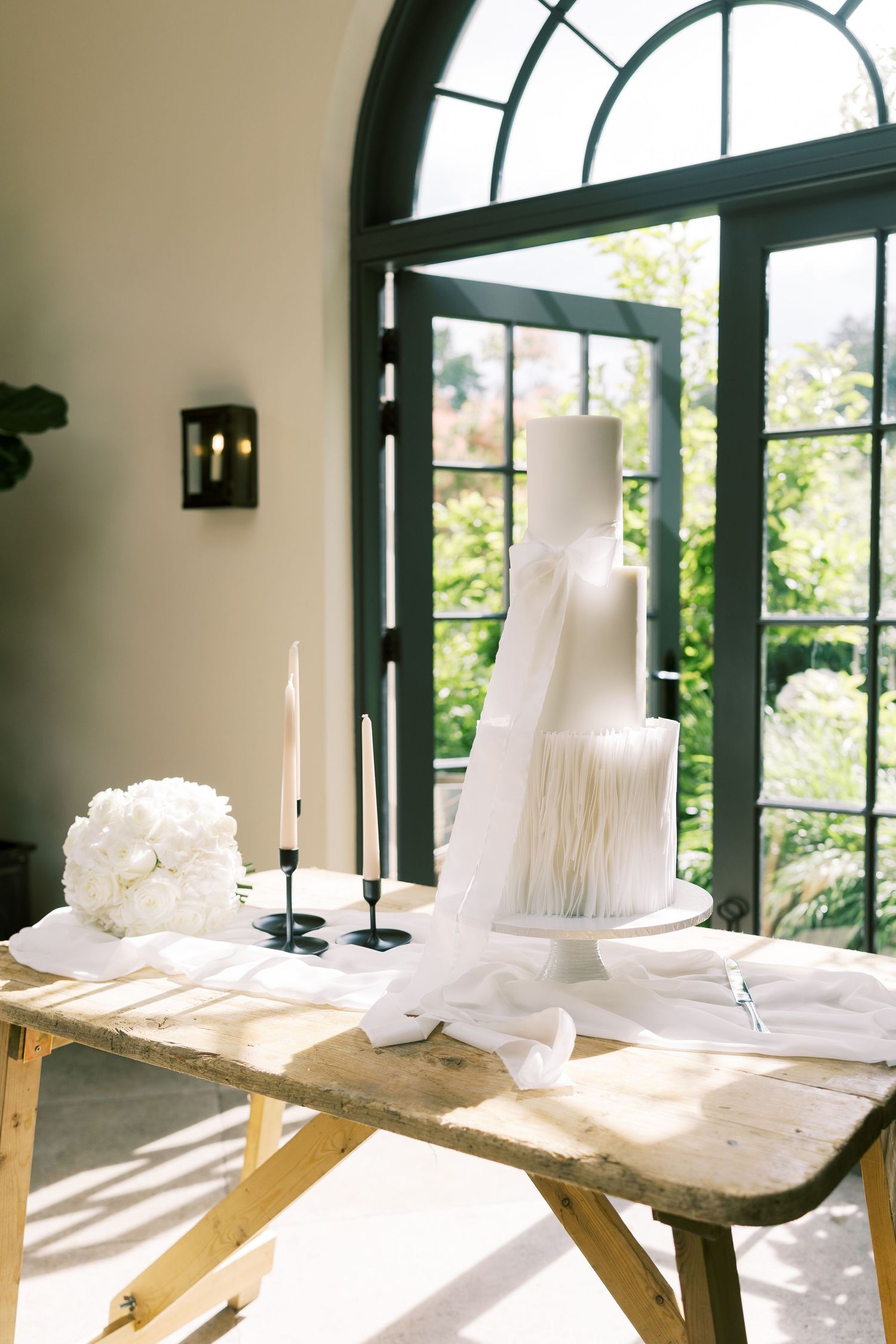 Elegant white tiered wedding cake with fringe details displayed on a rustic wooden table beside candles and floral bouquet.