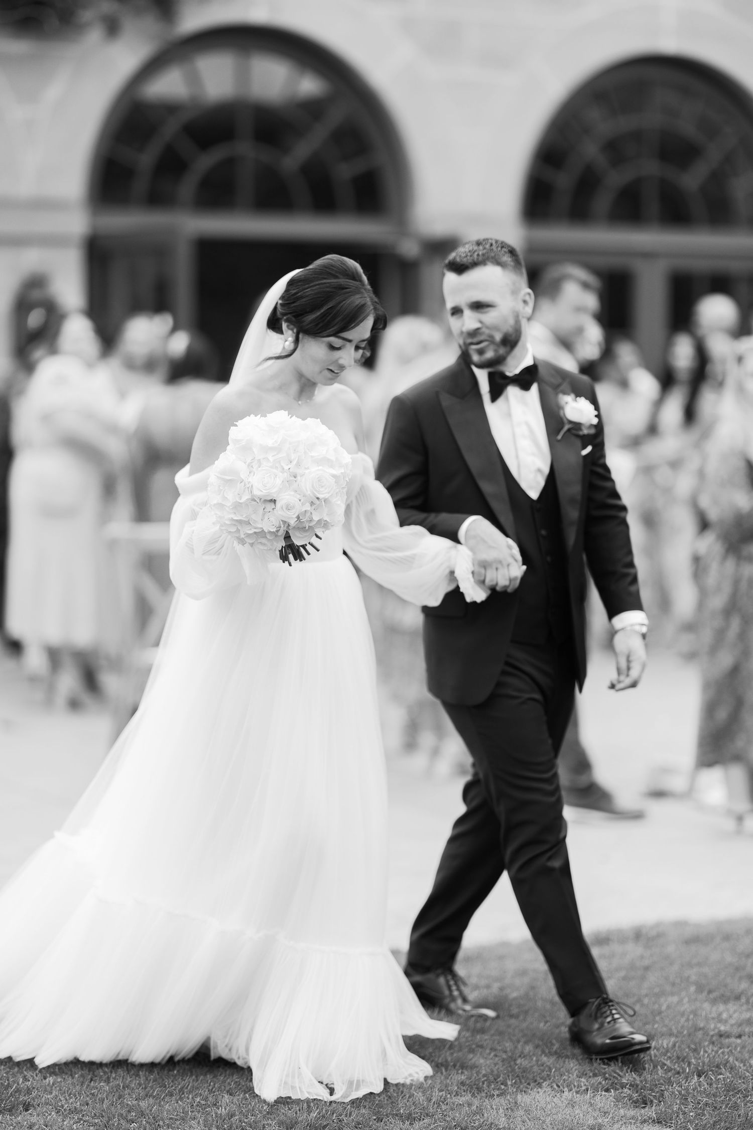 Black and white photo of elegant bride and groom walking hand in hand outdoors at a classic wedding venue.