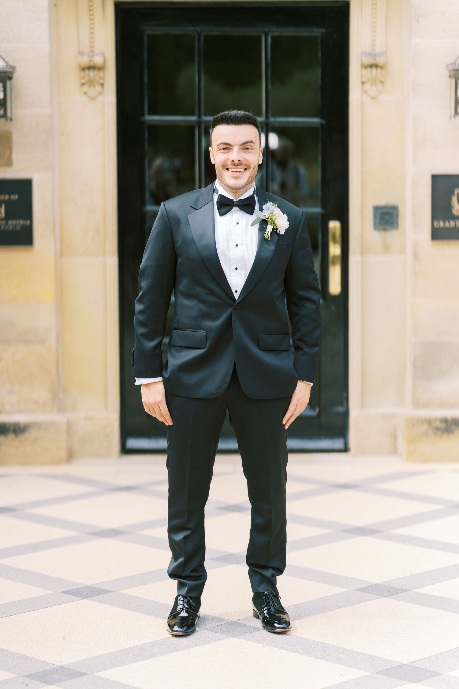 Smiling groom in black tuxedo with boutonniere standing outside elegant building entrance.
