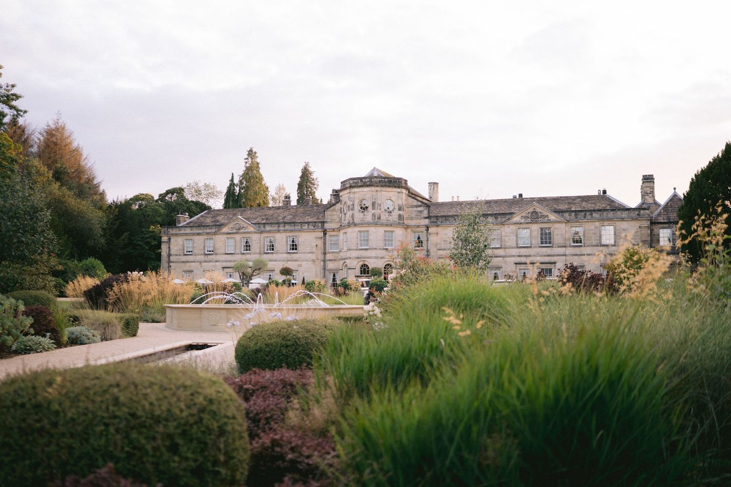 Elegant stone country house with manicured gardens, ornamental hedges, and autumn trees under a cloudy sky.