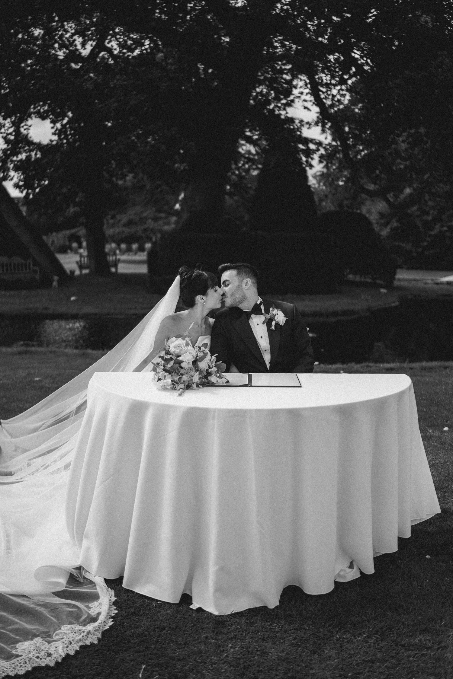 Black and white photo of bride and groom sharing a kiss at signing table with floral bouquet outdoors.