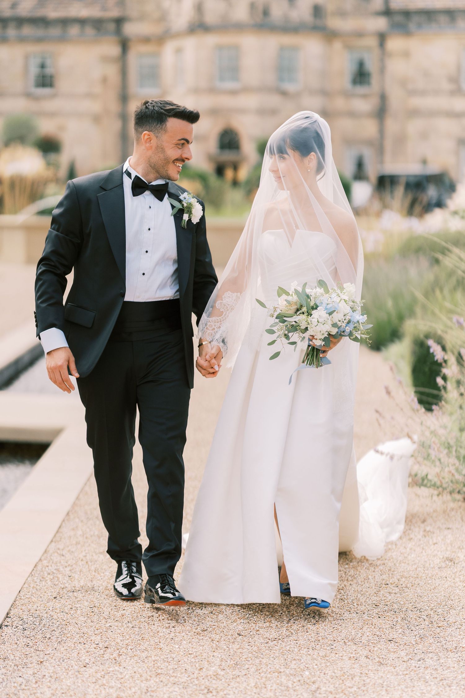 Bride and groom walk hand in hand outdoors, she in a white gown with veil, he in a black tuxedo with bow tie.