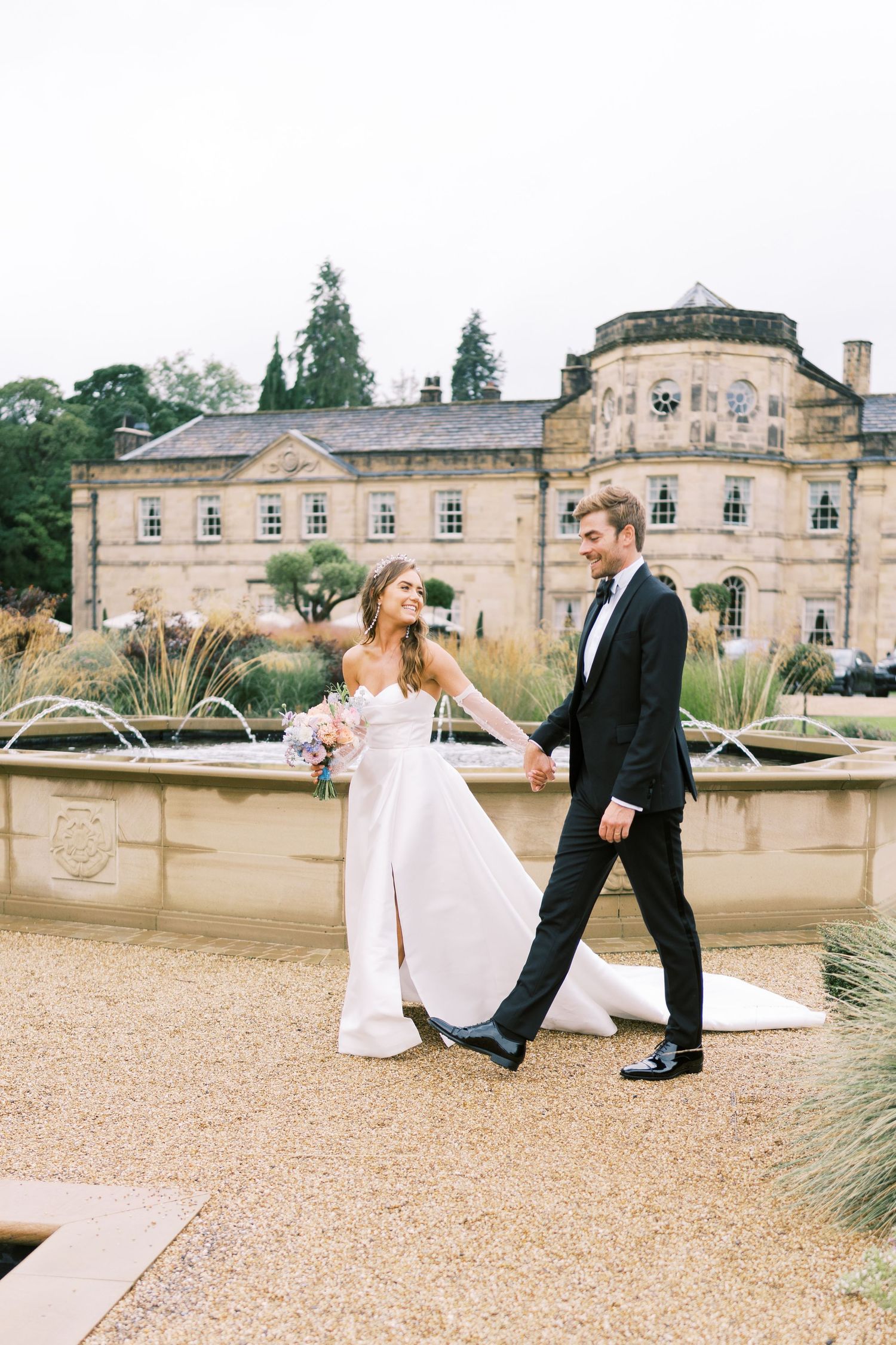 Bride and groom walk hand-in-hand before a grand stone manor fountain, she in a white gown, he in a black tuxedo.